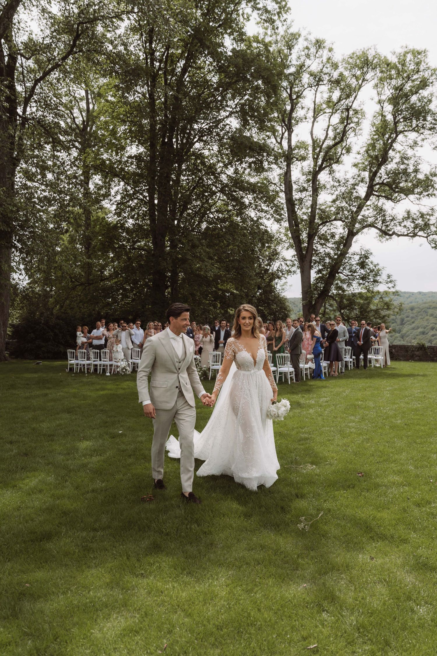 A couple walks across a lush green lawn after their outdoor wedding ceremony with guests in the background.