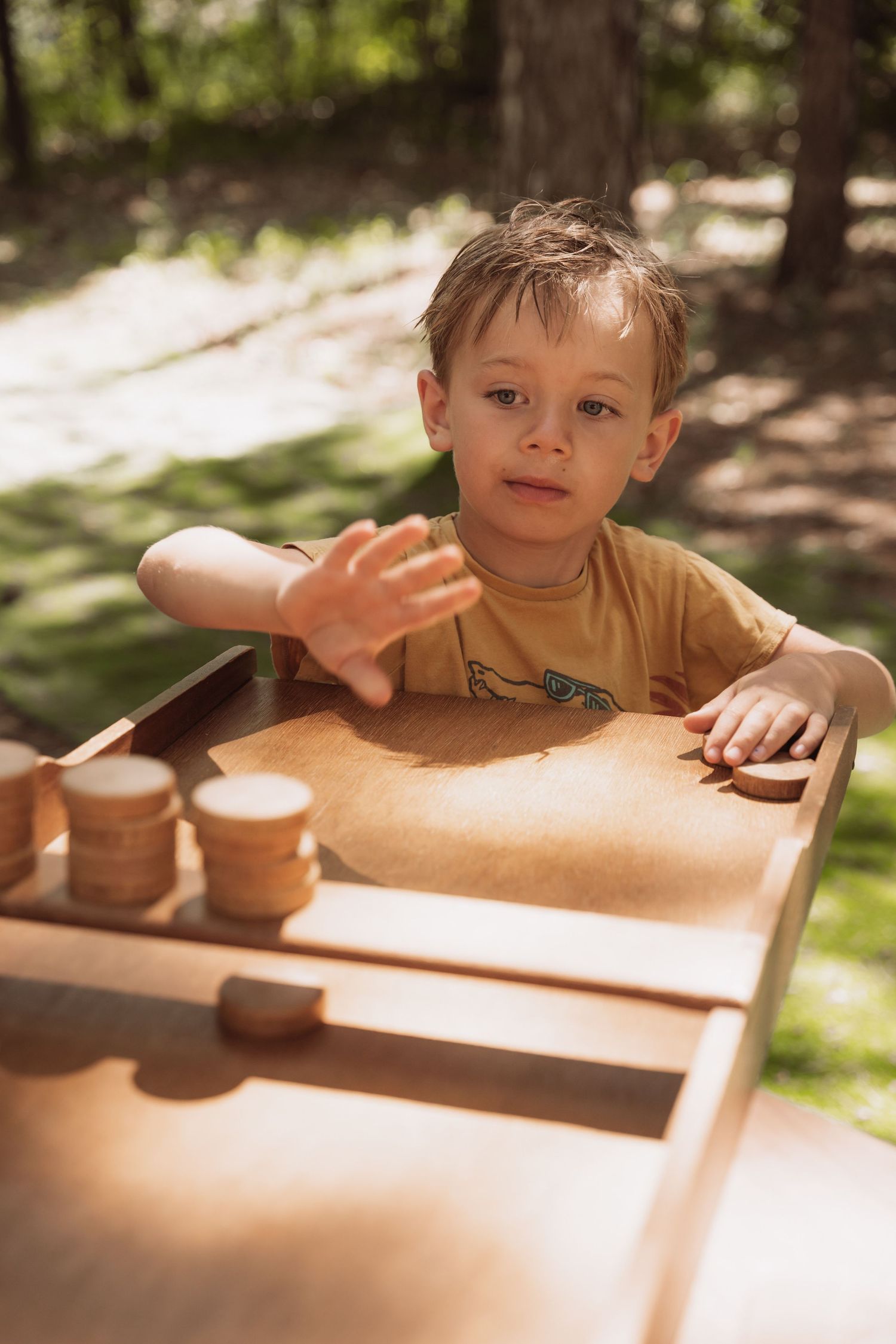 A child plays at a wooden table outdoors in dappled sunlight on a summer day.