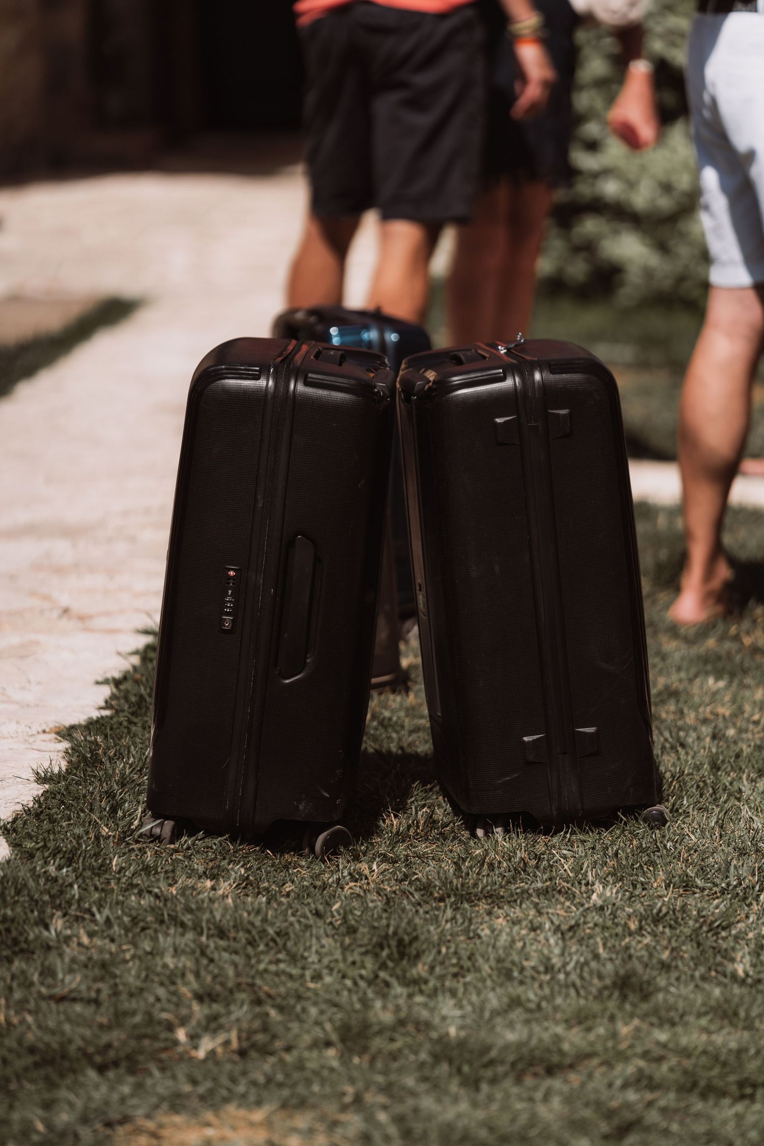 Two black hardshell rolling suitcases sitting on grass with people's legs visible in the background.