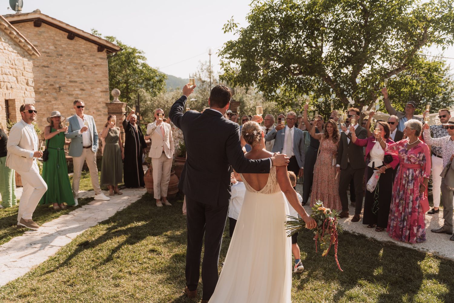 Wedding guests gather outdoors at a rustic venue with stone walls and greenery for a celebration ceremony.