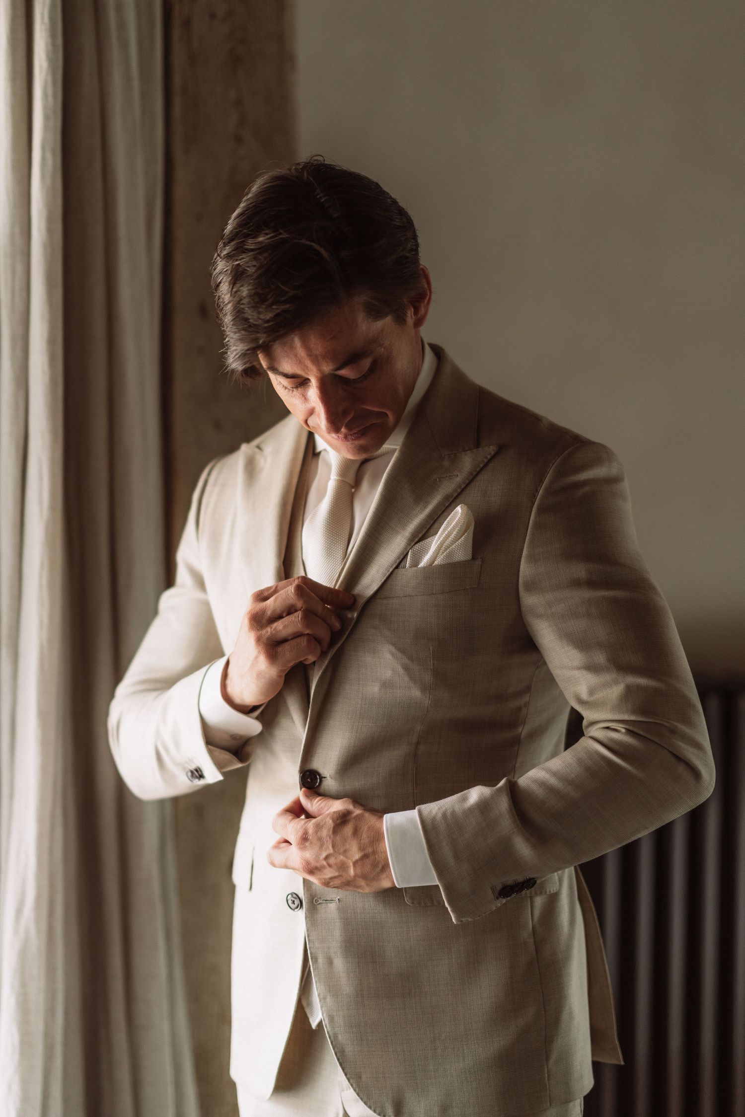 A groom adjusts his tie while getting ready in a beige suit by a window.