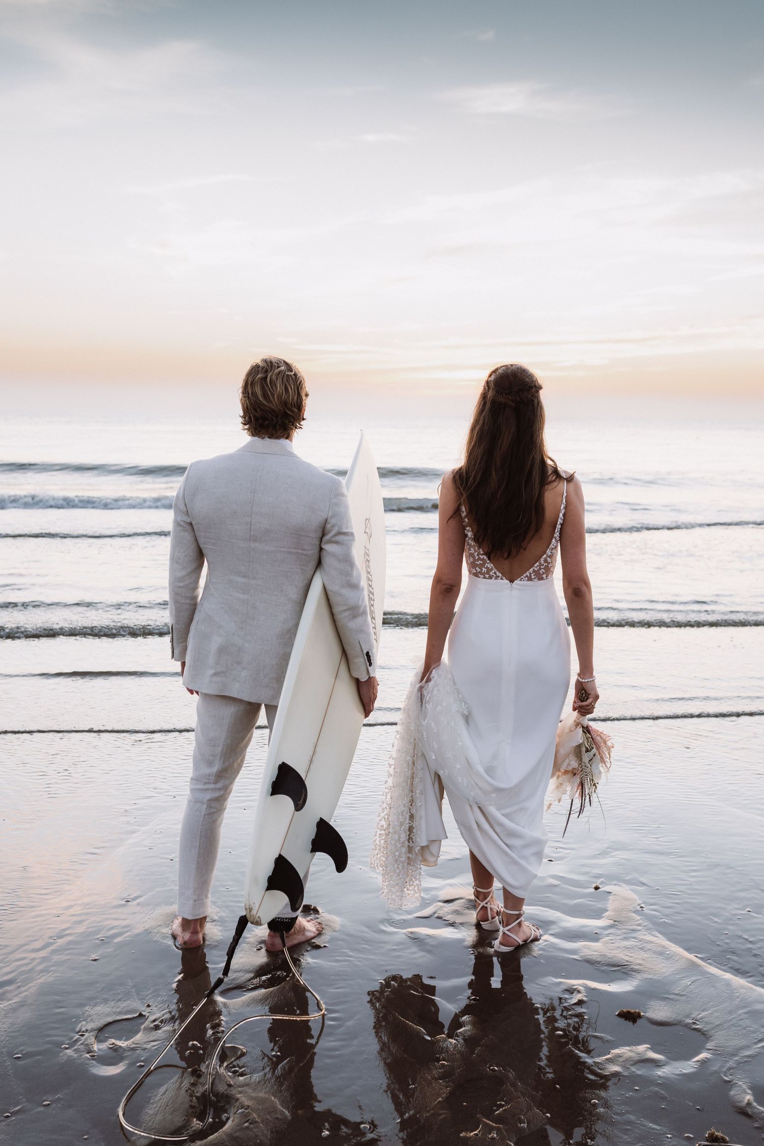 Couple in white attire walking along a beach at sunset holding a surfboard.