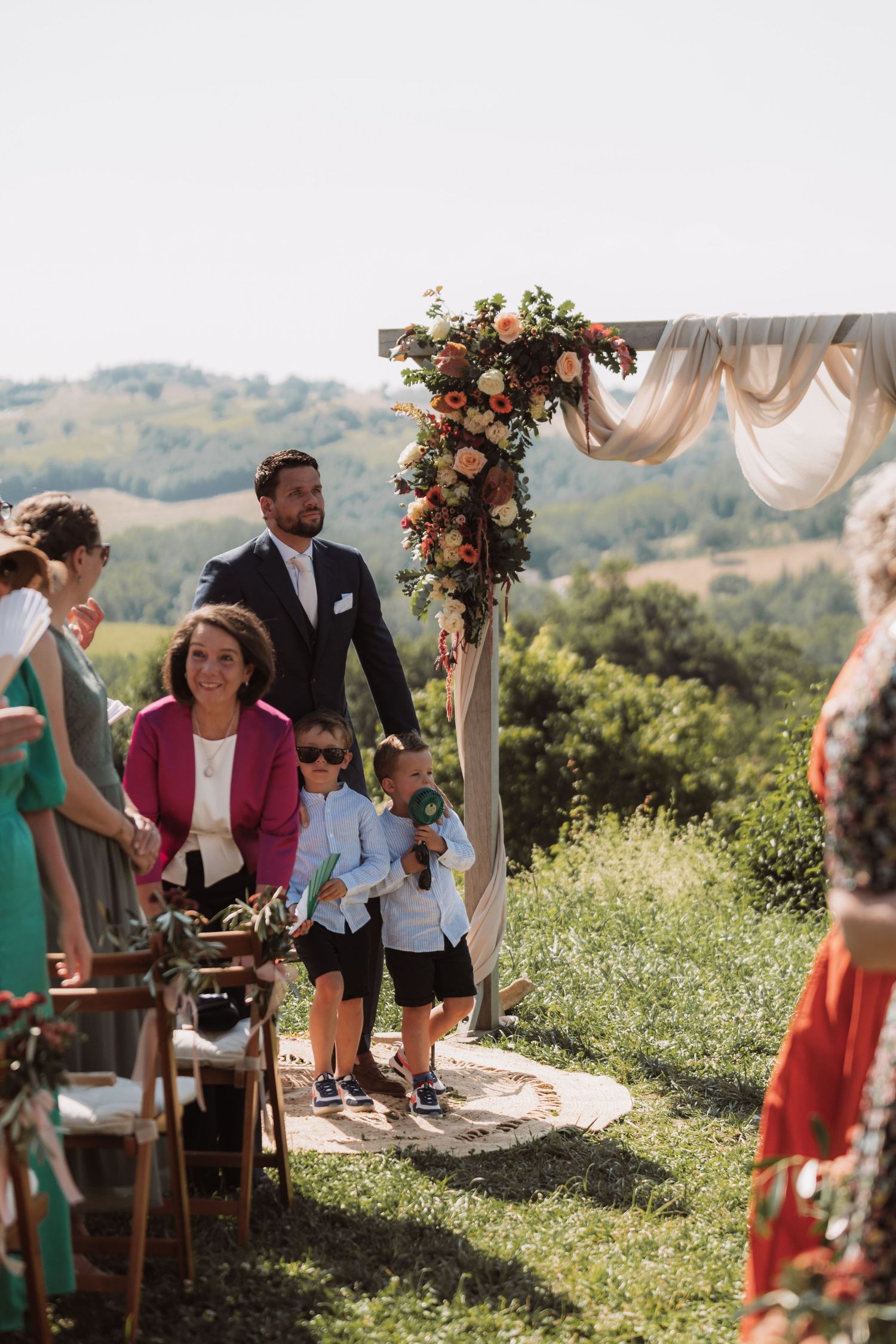 Outdoor wedding ceremony on a hilltop with floral arch decorations and guests gathered on a sunny day.