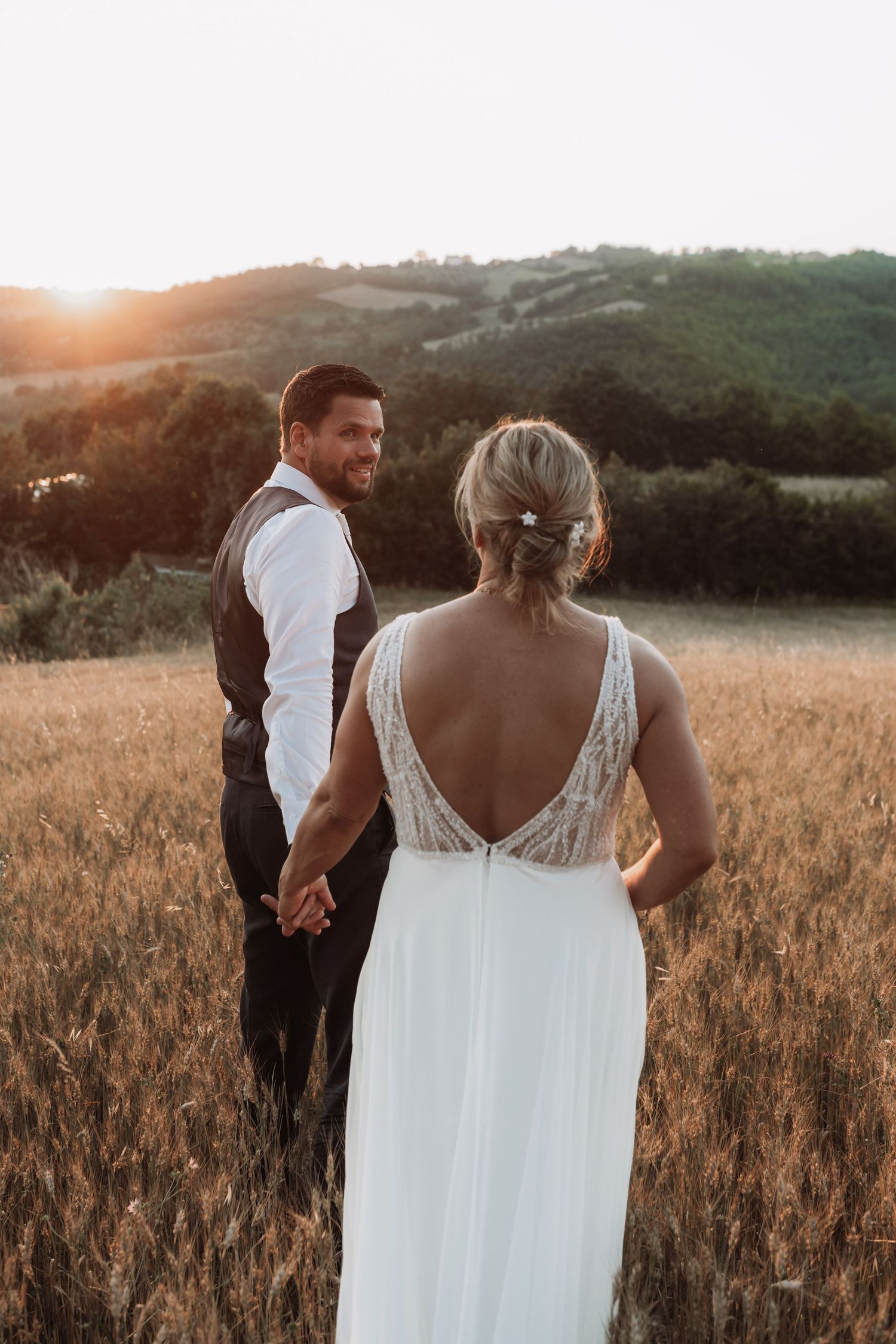 Couple holding hands in golden field at sunset with mountains in background.