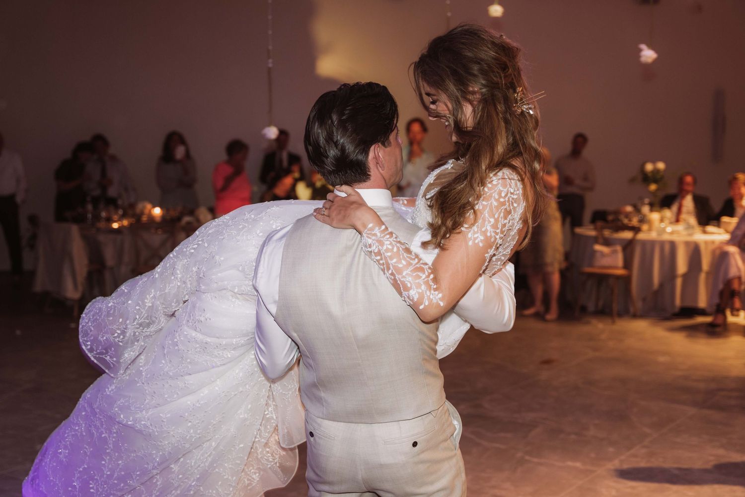 A couple shares their first dance at a wedding reception with soft purple lighting and guests in the background.