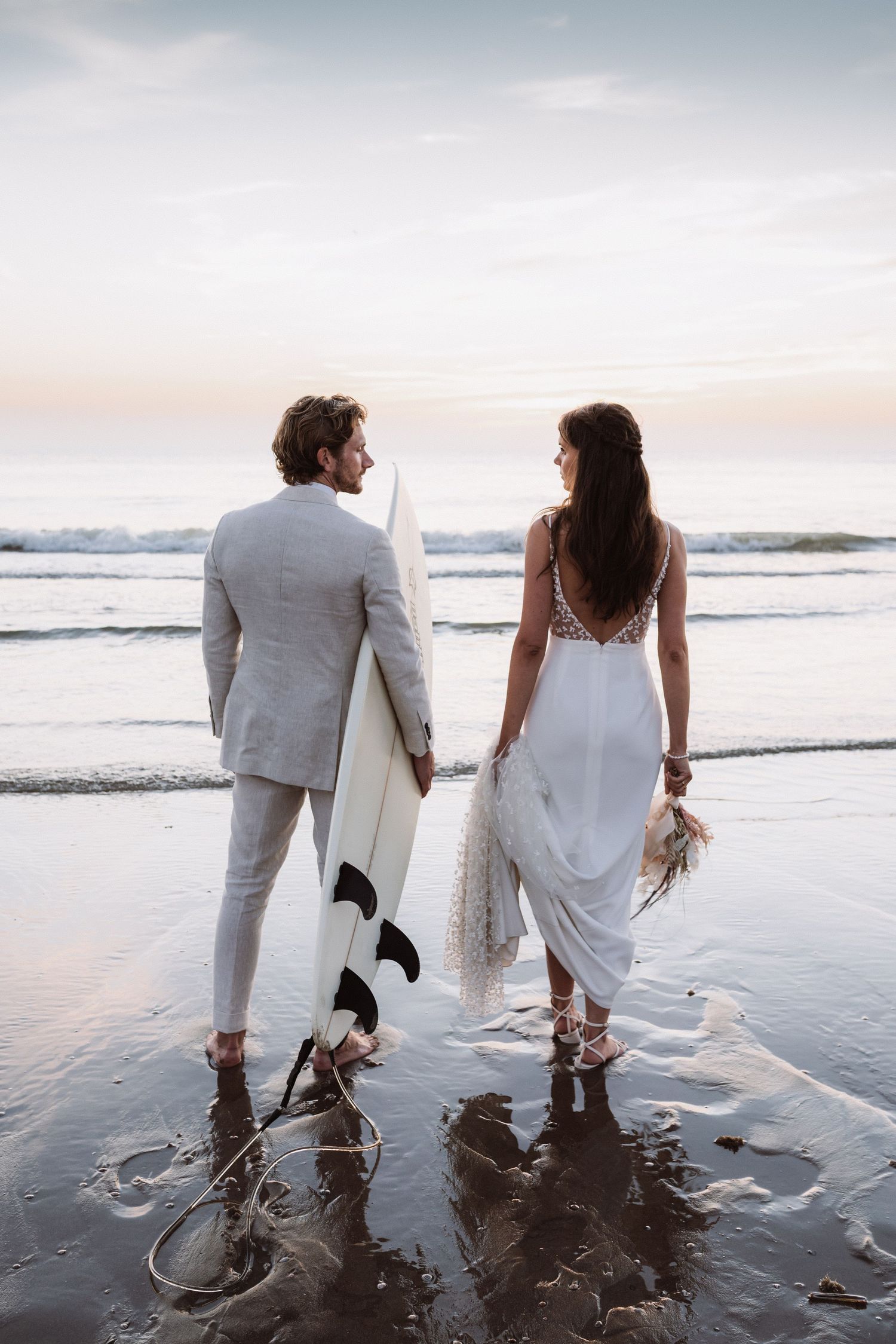 Couple standing in shallow ocean water with a surfboard during sunset.