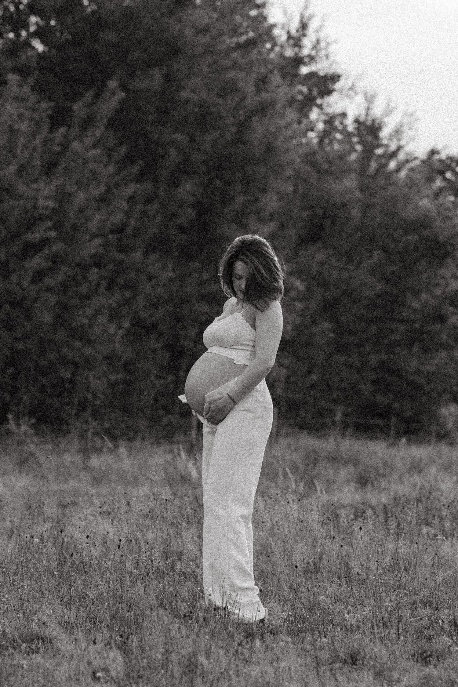 Black and white maternity photos in flowing white dress in grassy field.
