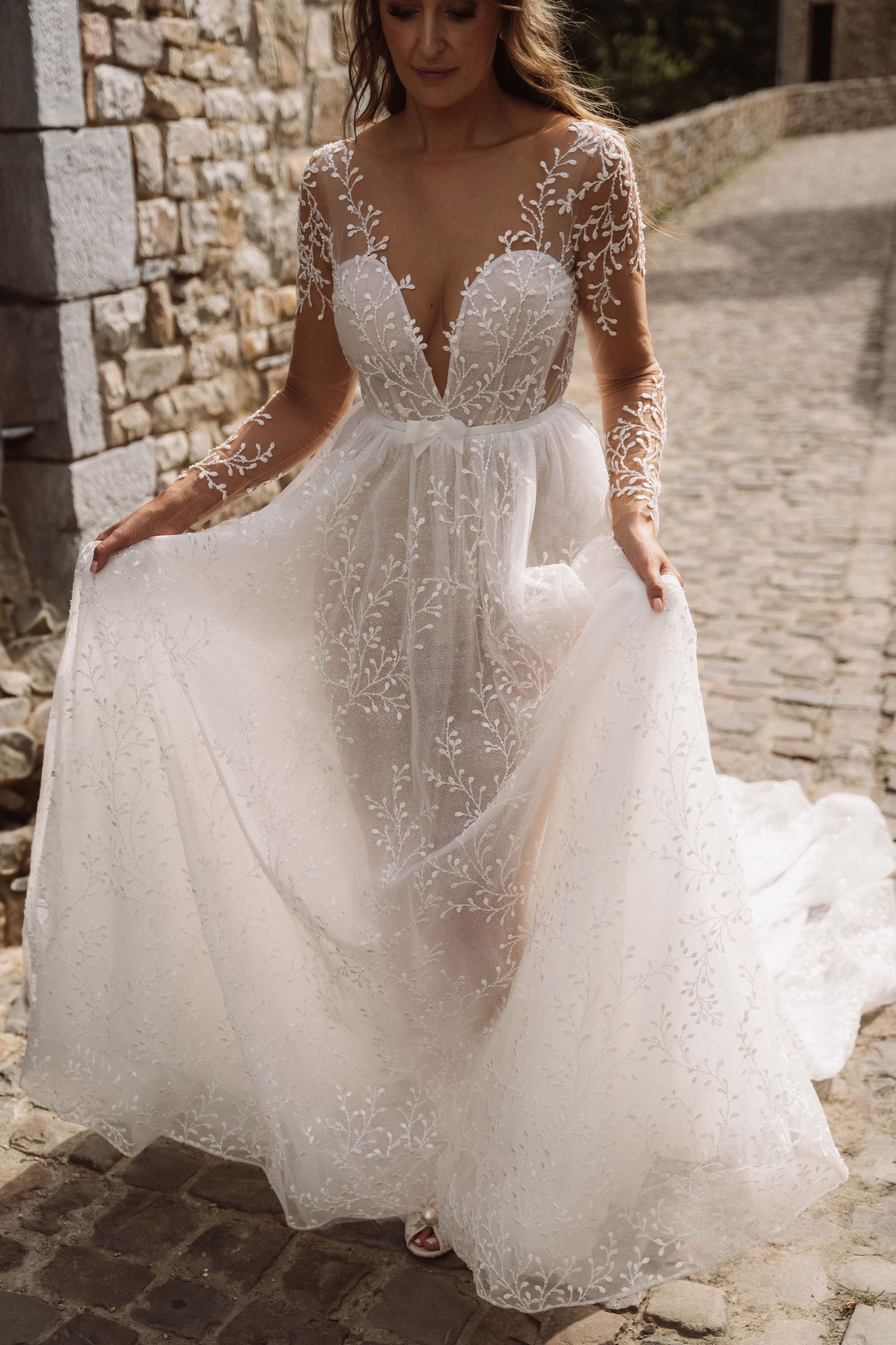 A flowing white wedding dress with delicate lace details and sheer long sleeves shown in multiple angles on a stone path.