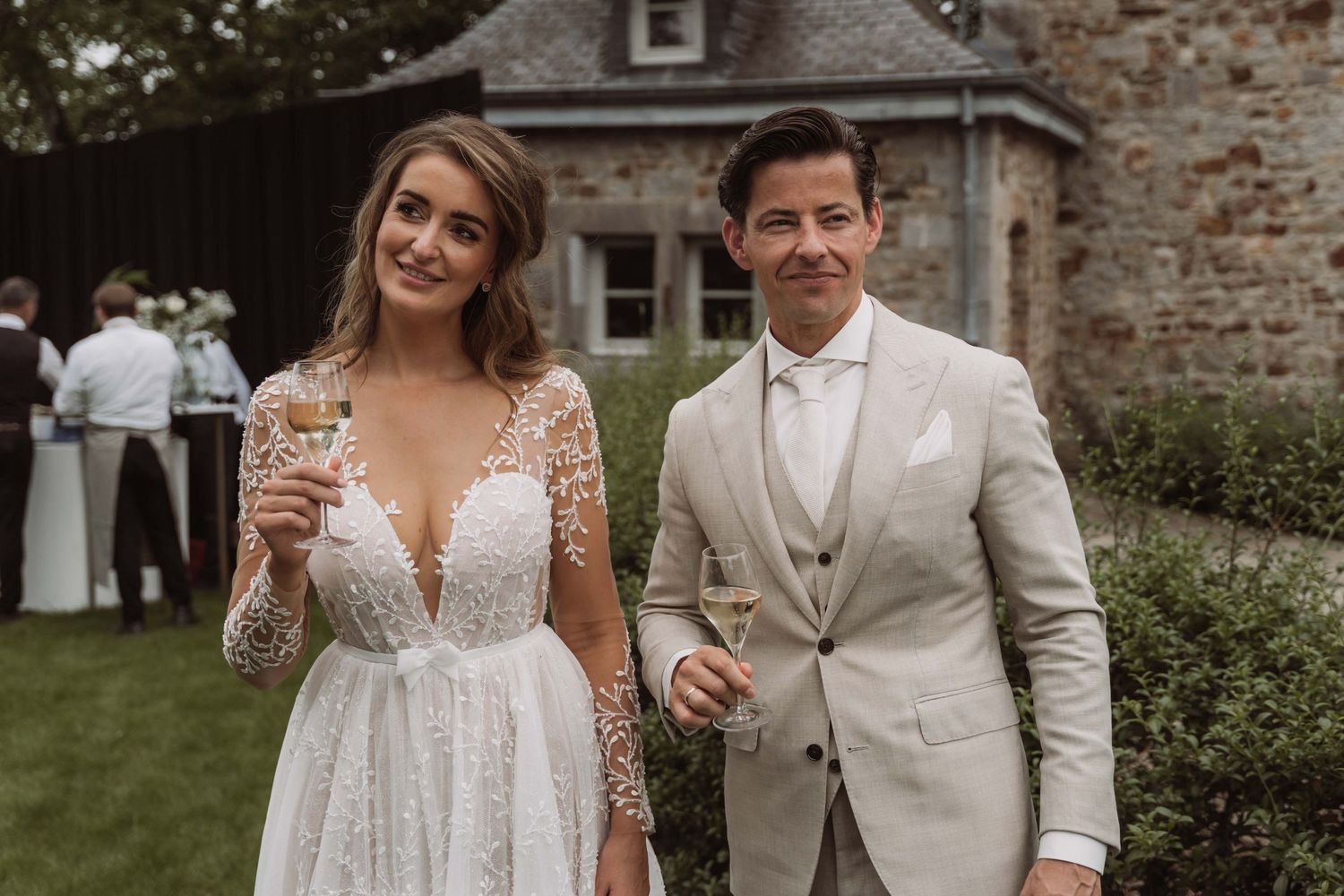 A wedding couple shares a celebratory toast outdoors near a rustic brick building.