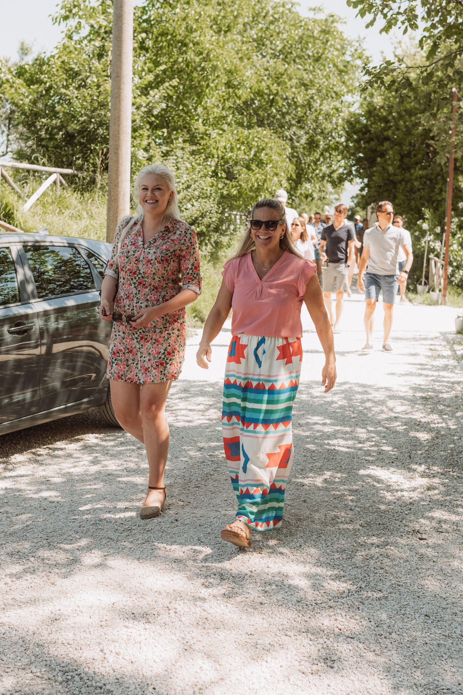 People walking on a gravel path through a sunny park area carrying colorful beach accessories.