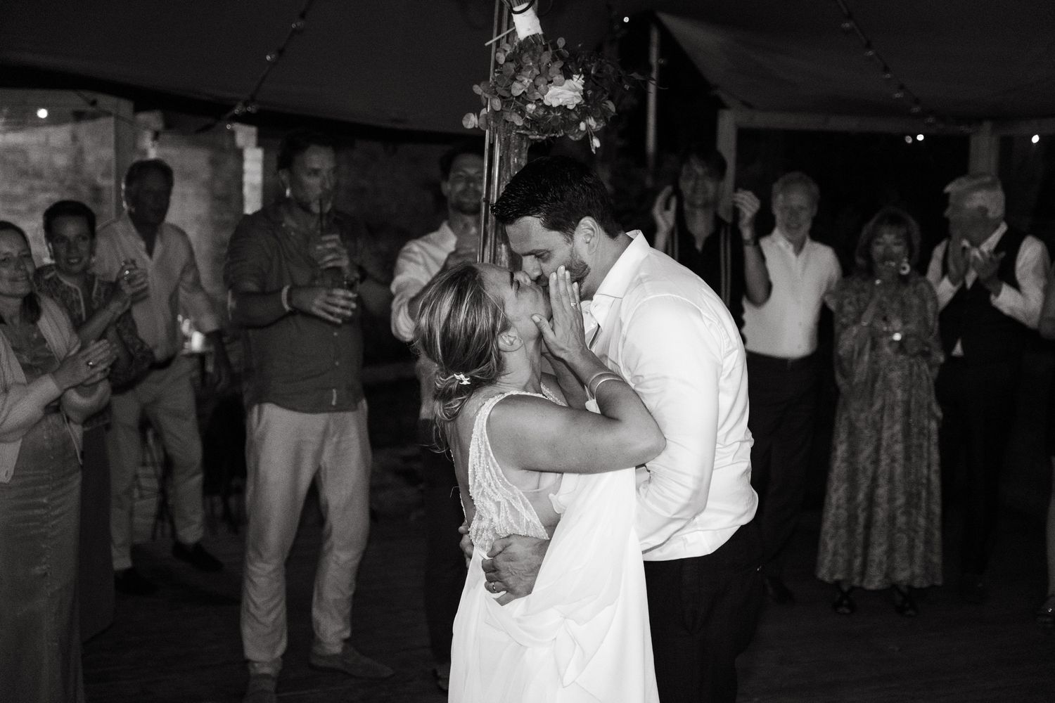 A couple shares their first dance at a wedding reception while guests watch from the sidelines in black and white photography.