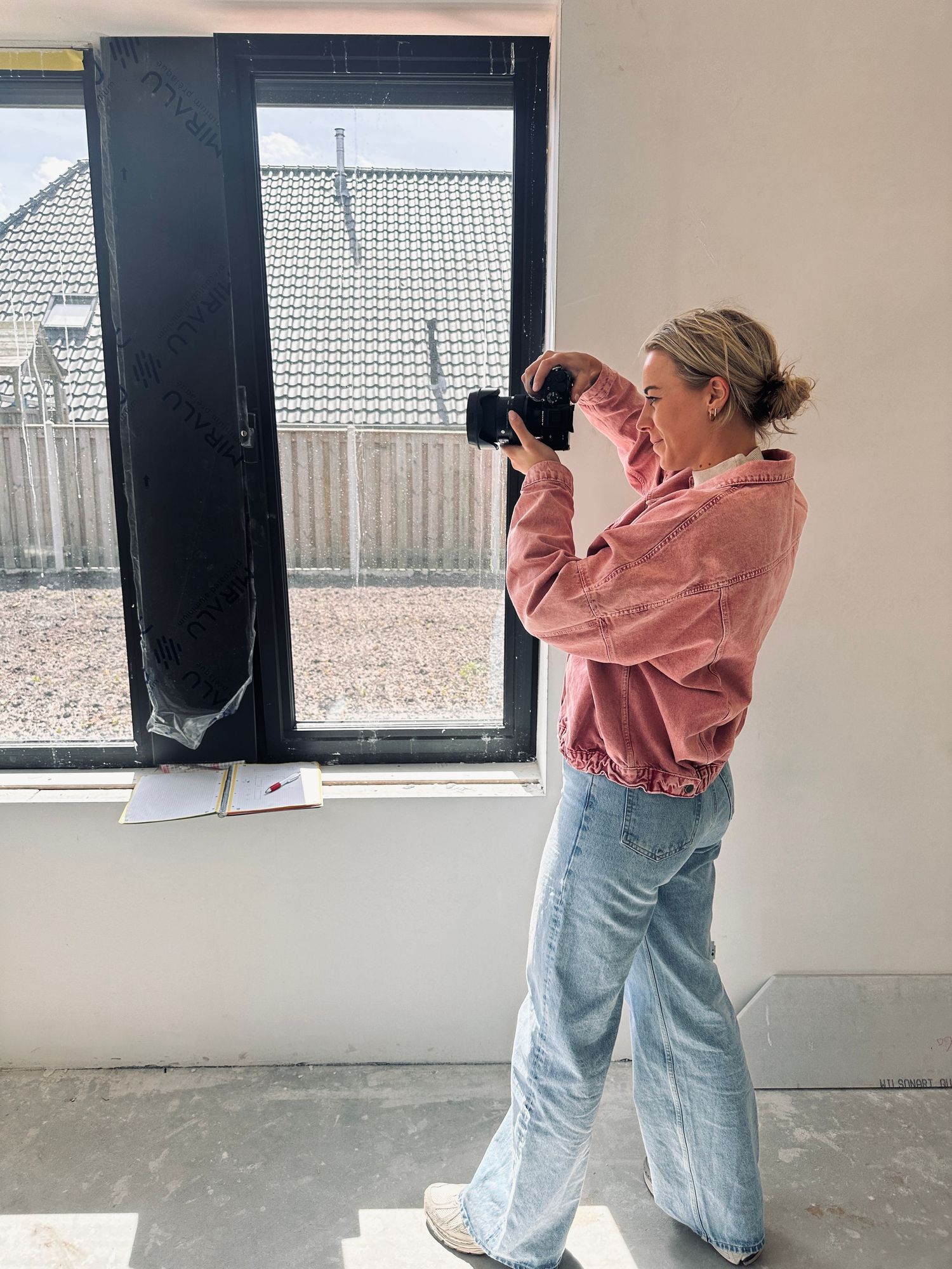 Photographer in pink hoodie and jeans taking pictures near a window.