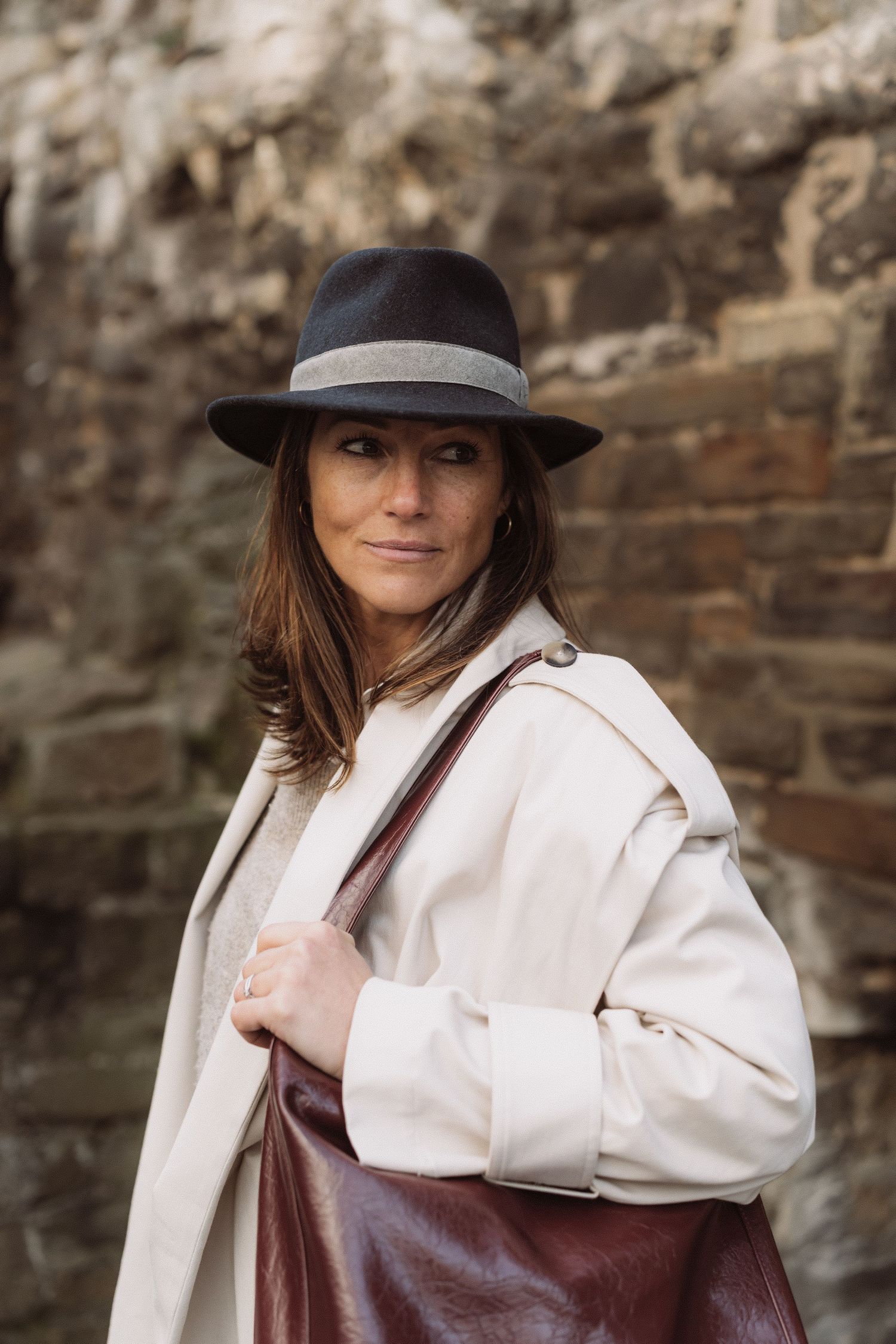 A person wearing a wide-brimmed hat and cream coat poses against a stone wall with a brown leather bag.