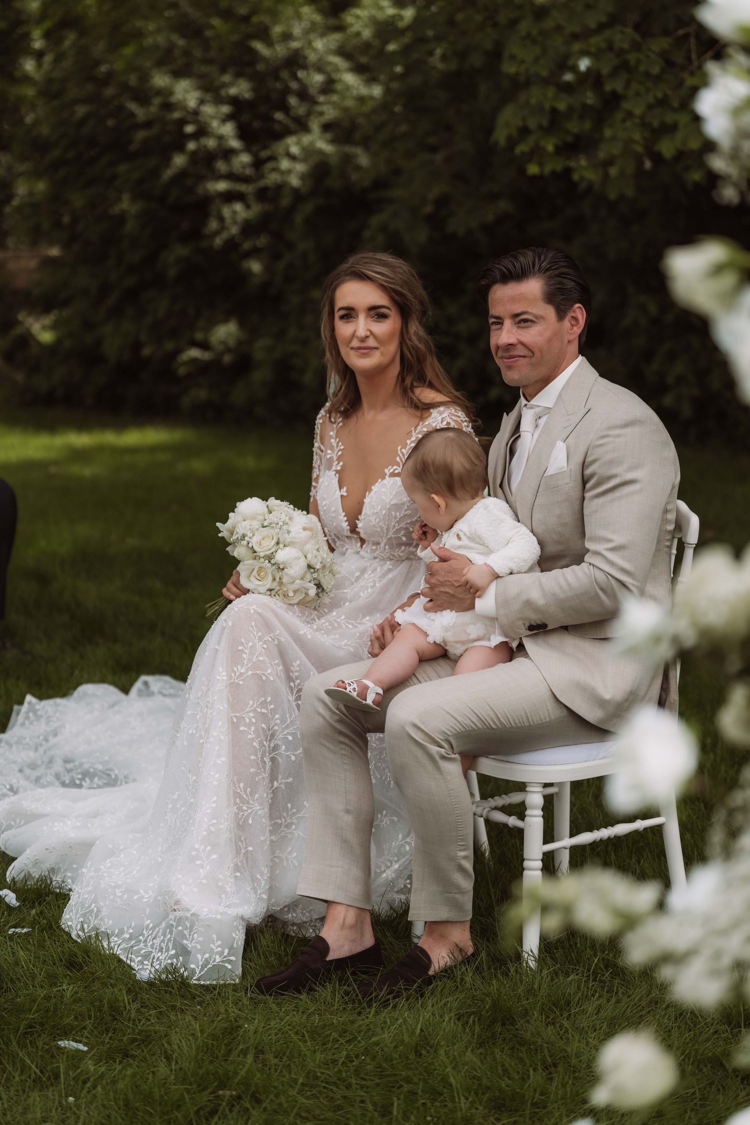 A couple in wedding attire sits together on white chairs in a lush garden setting surrounded by flowering trees.