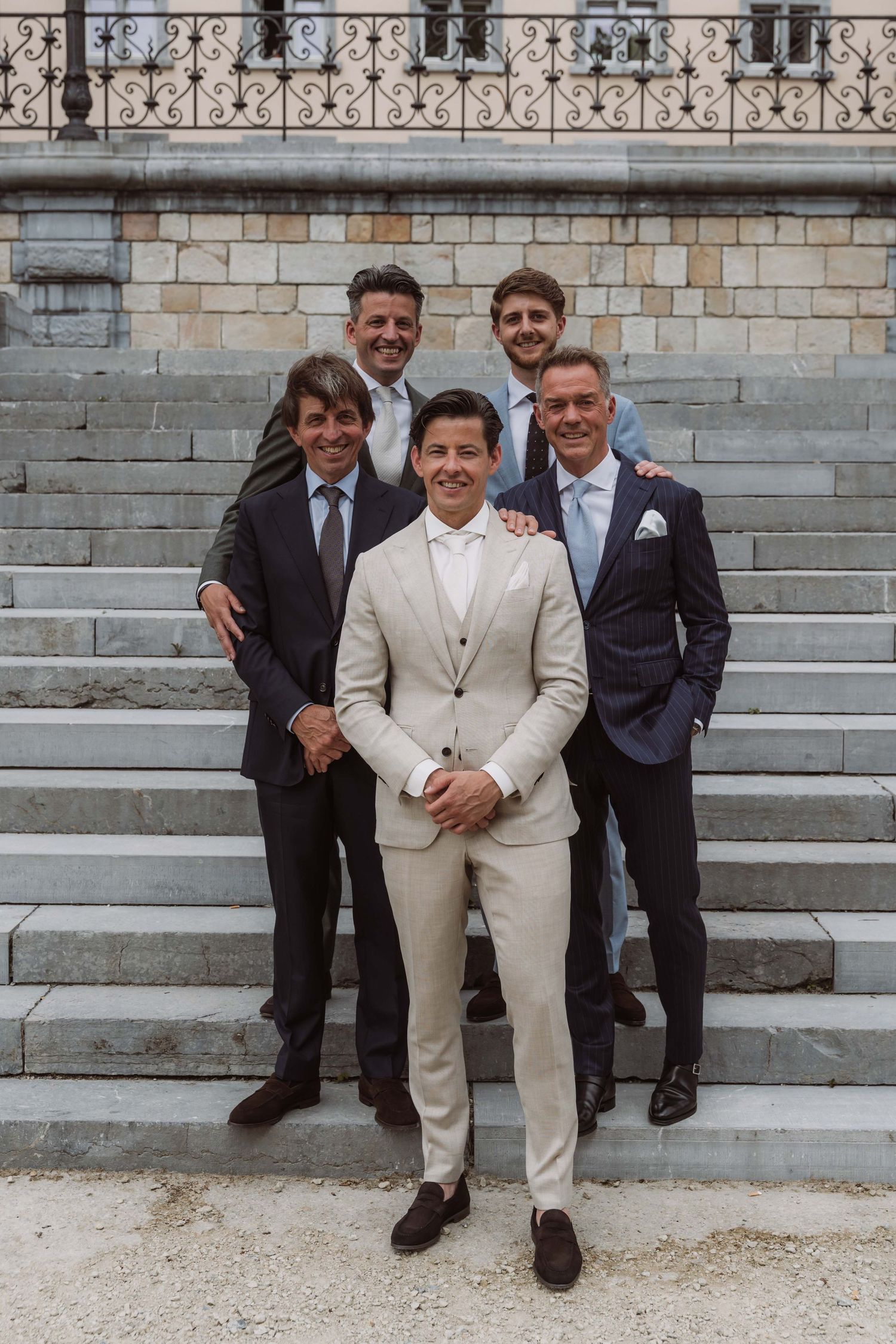 Three people in formal attire sitting on stone steps, with one wearing a light colored suit and two in dark suits.