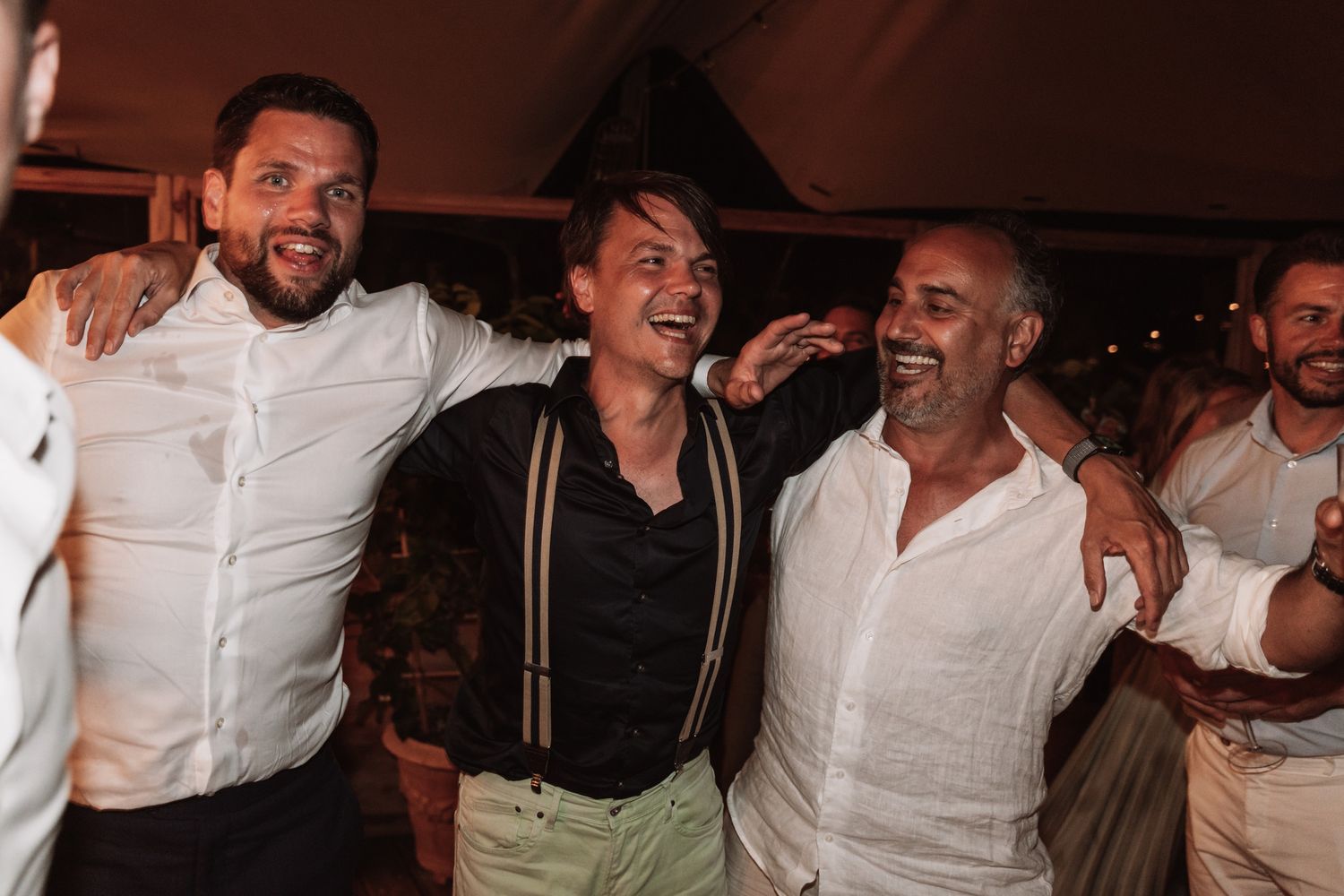 Three people smiling and embracing at a party, wearing white shirts and suspenders in a dimly lit venue.