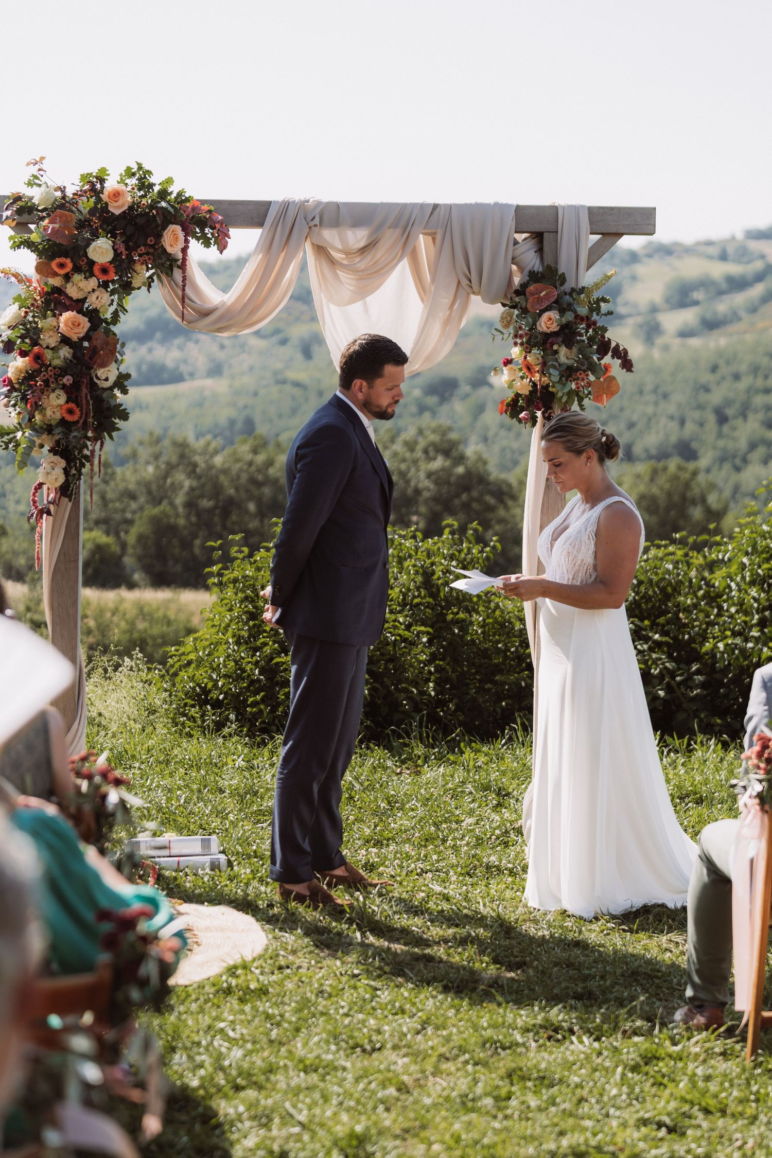Outdoor wedding ceremony with couple exchanging vows under floral arch decorated with blush drapery on hilltop.