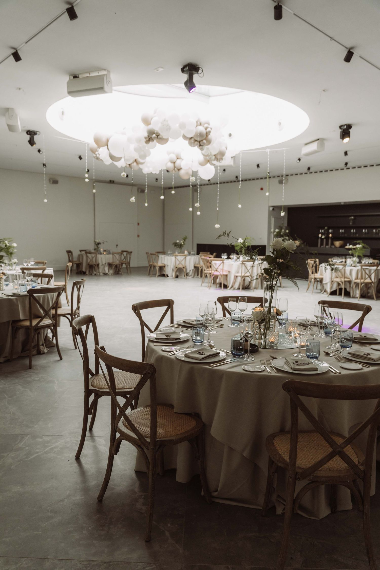 Elegant wedding reception venue setup with round tables, wooden cross-back chairs, and ambient overhead lighting.