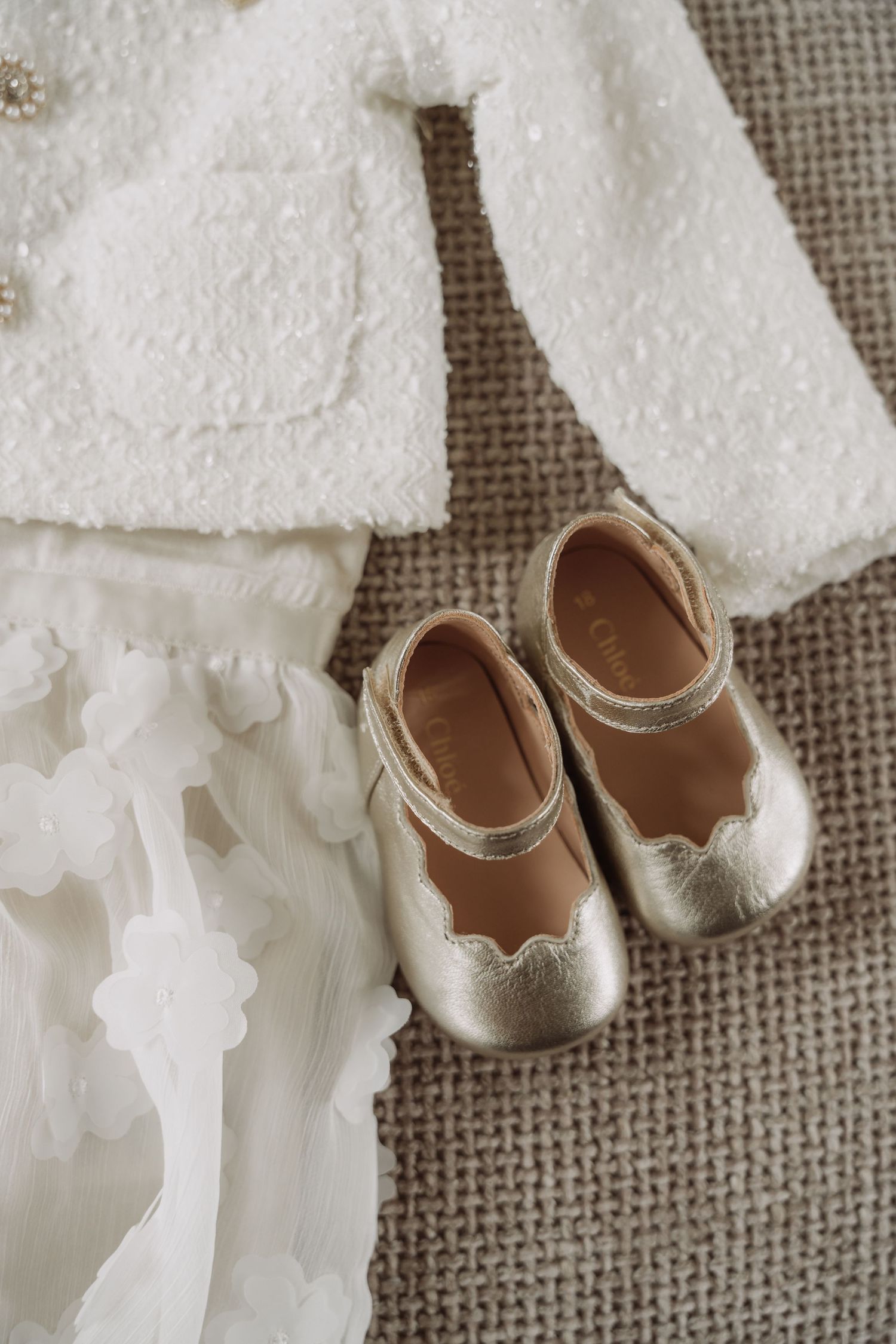Three pairs of delicate white baby shoes arranged in a row on a textured burlap background.