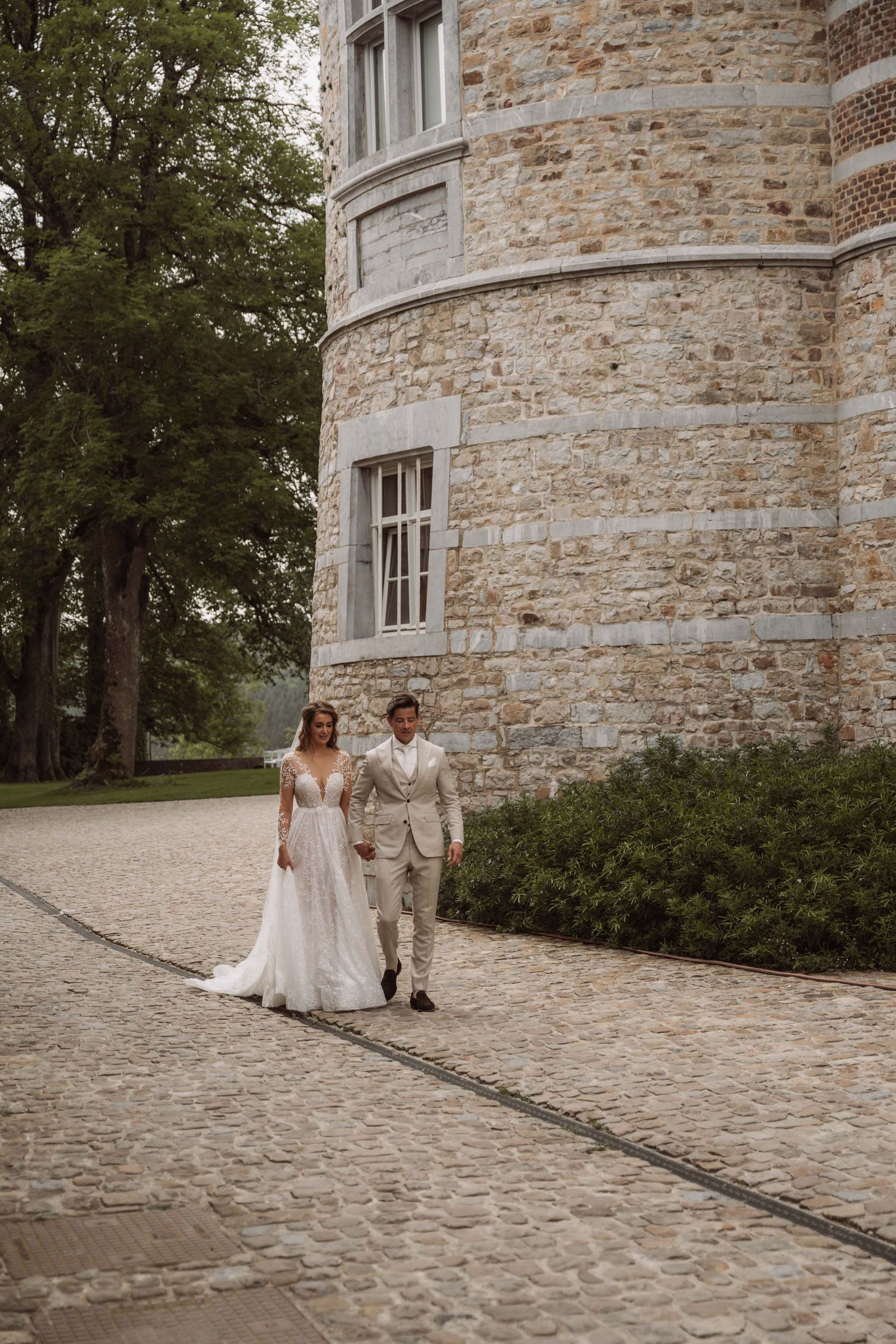A couple in wedding attire walks together along a curved stone path beside a rustic stone building exterior.