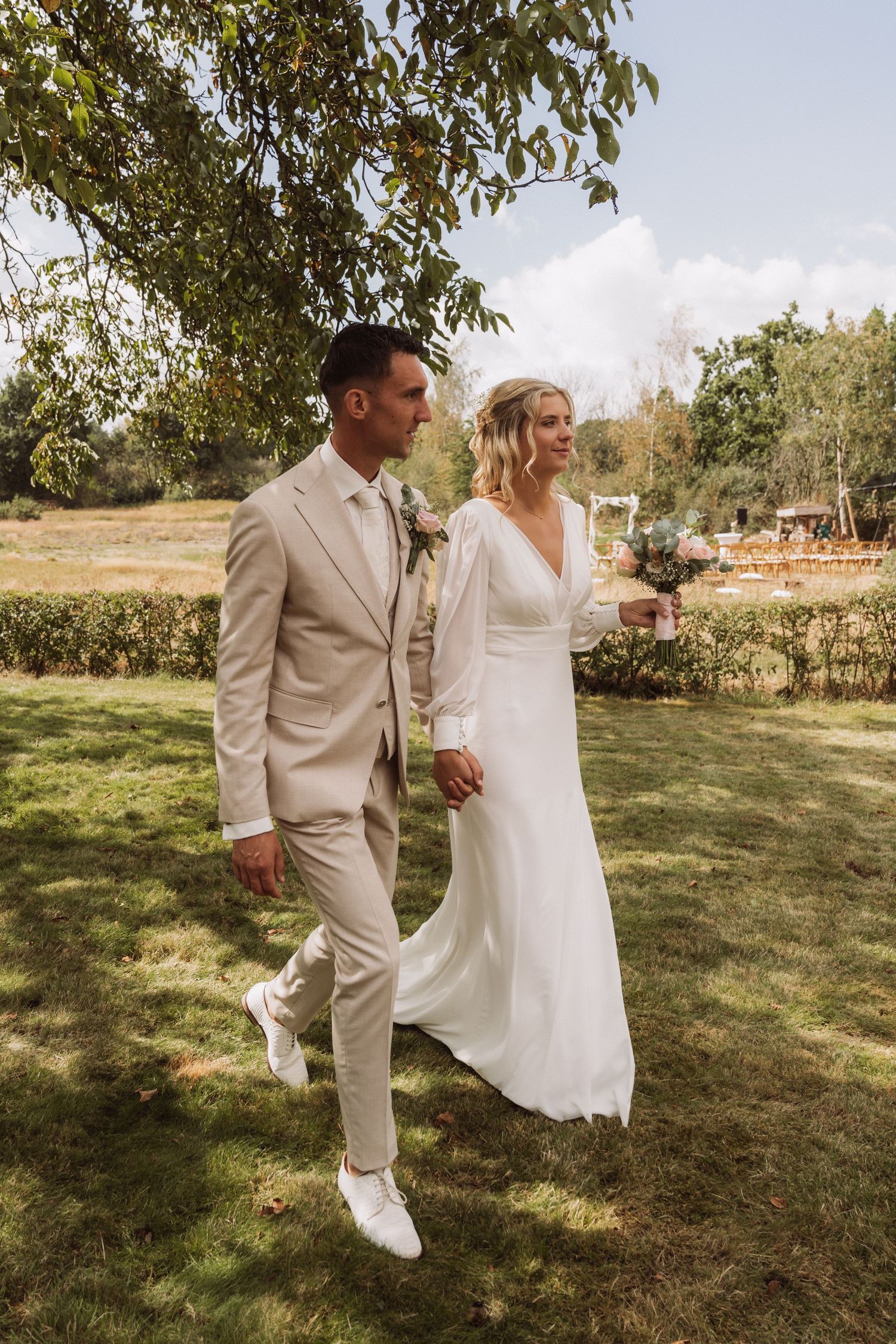 A couple in light colored attire walking through a sunny garden setting.