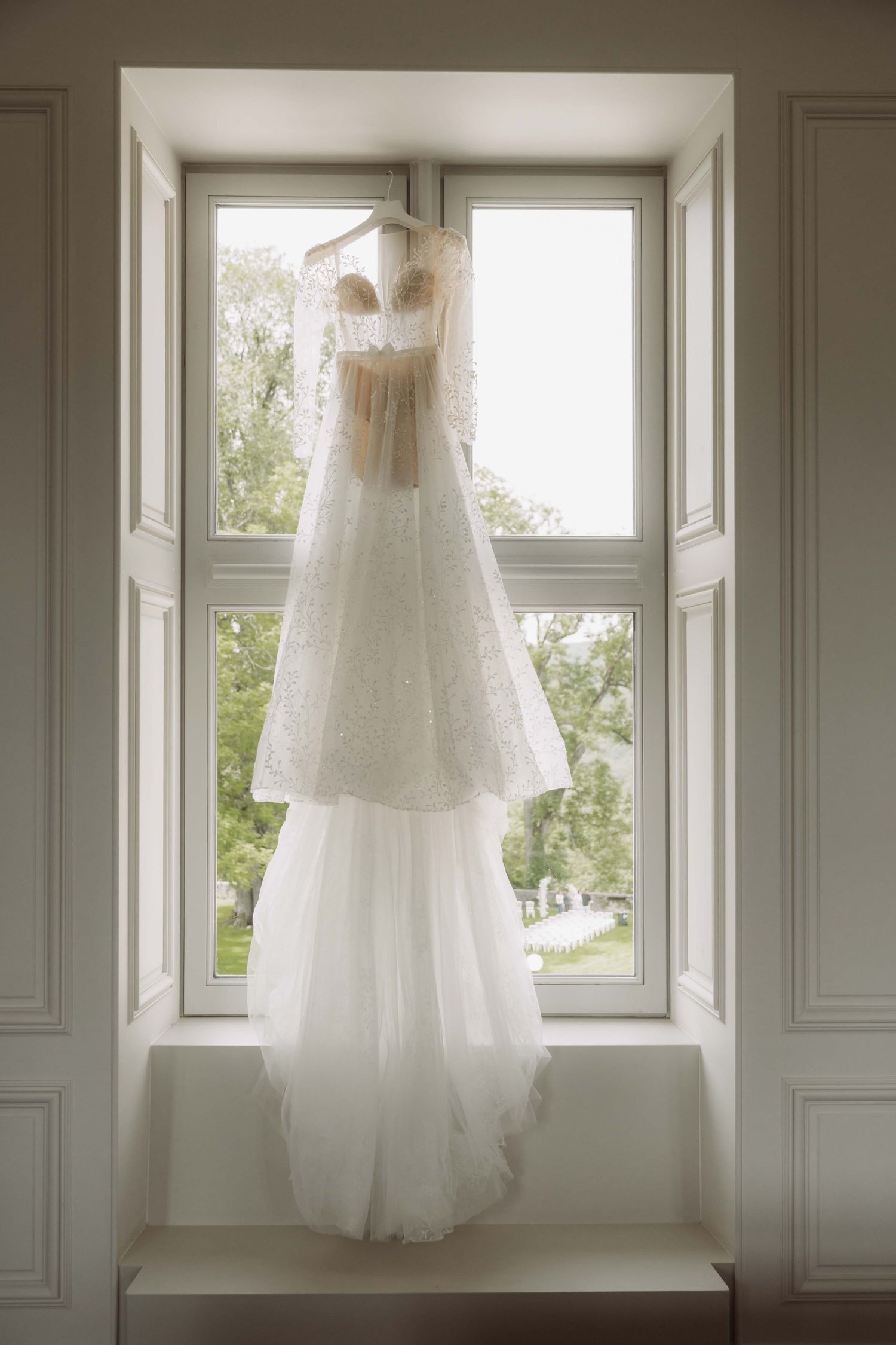 Three elegant white wedding dresses hanging in bright windows with natural light streaming through sheer fabric.