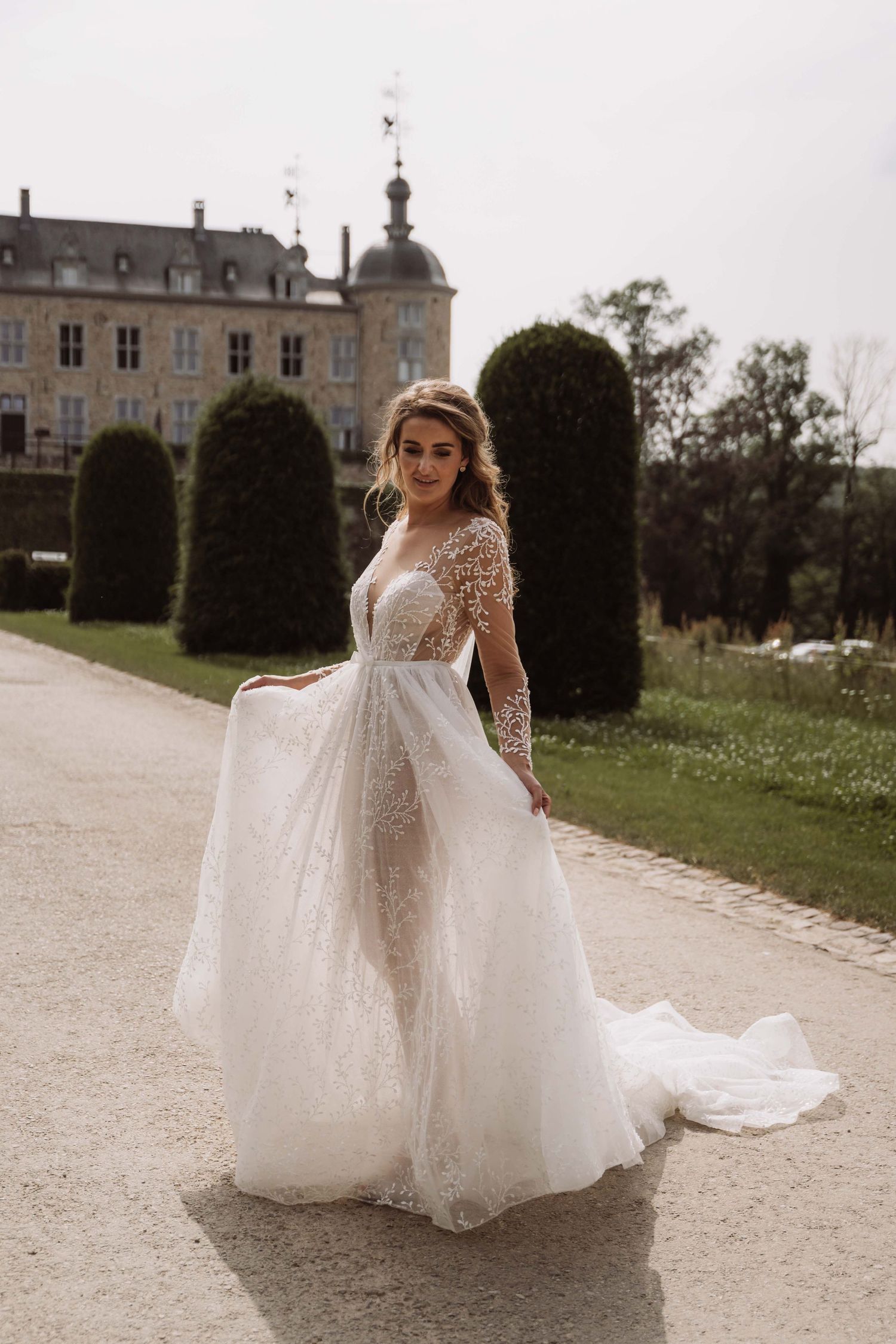 Bride in flowing lace wedding gown with long train walks along path outside elegant French chateau on sunny day.