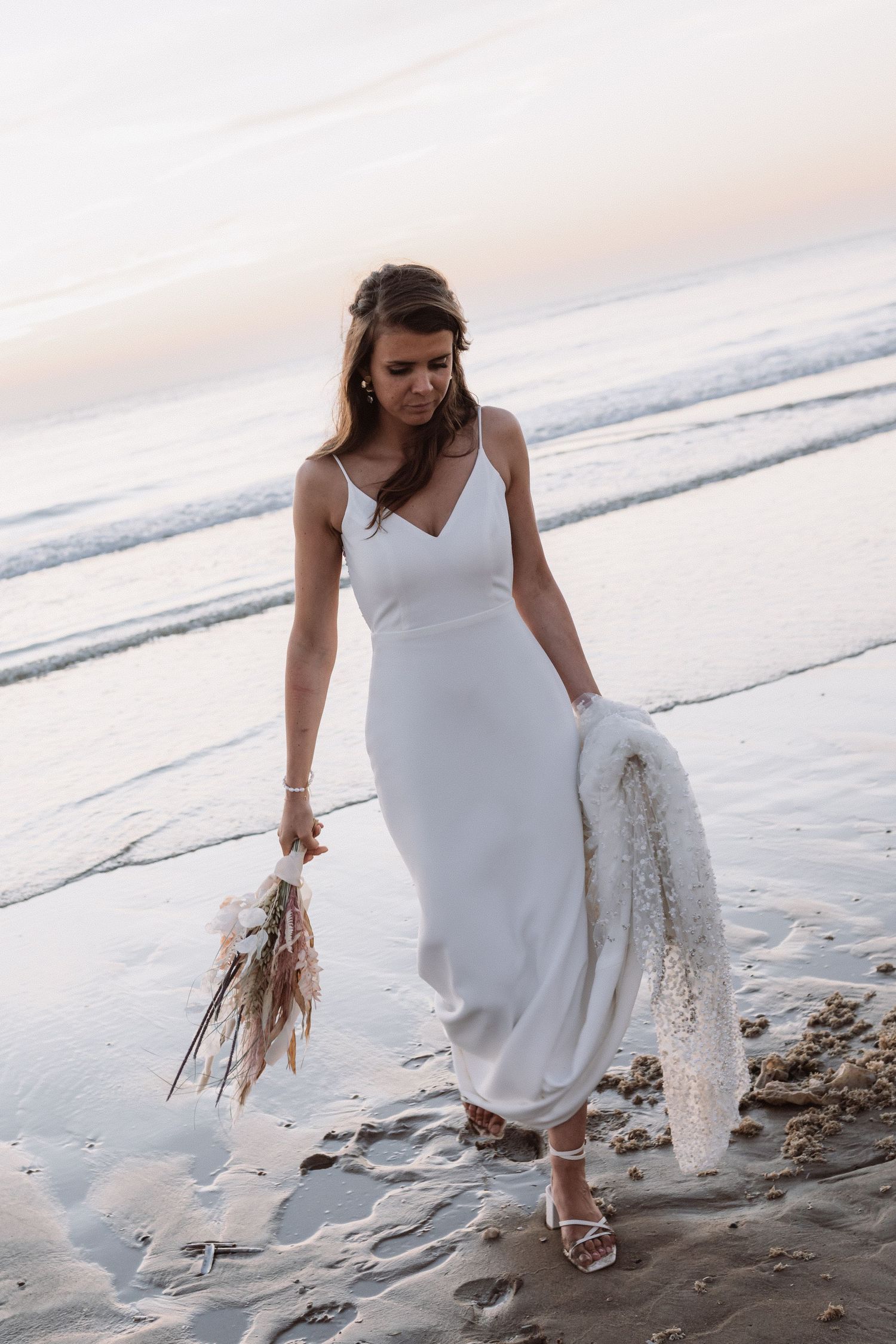 Woman in white dress walking along beach shoreline at sunset.