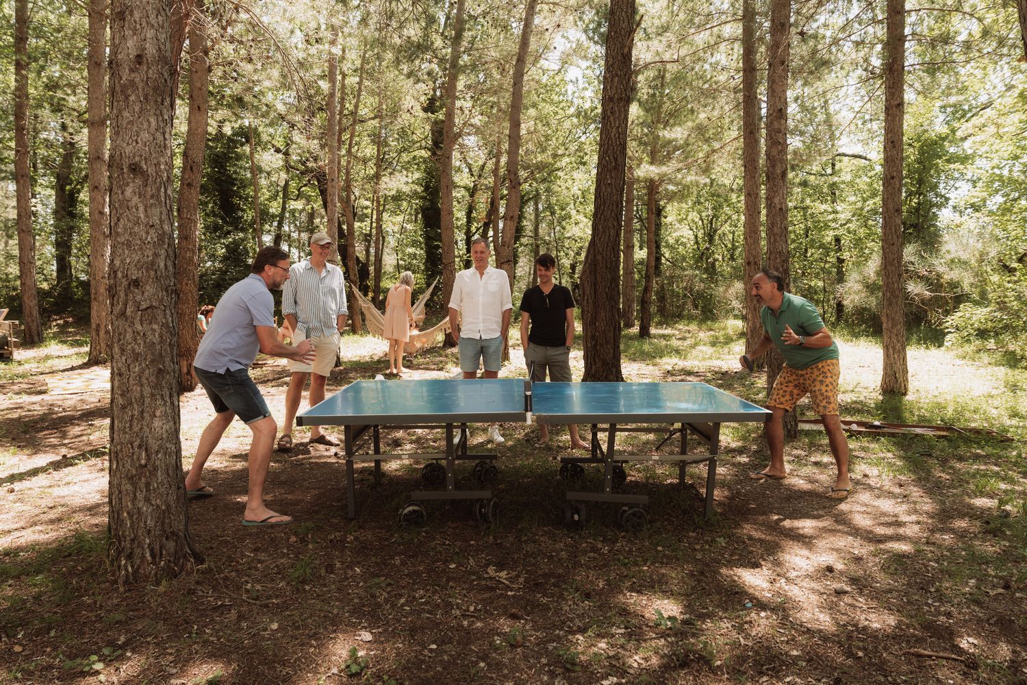 A group plays table tennis outdoors in a wooded area surrounded by tall pine trees on a sunny day.