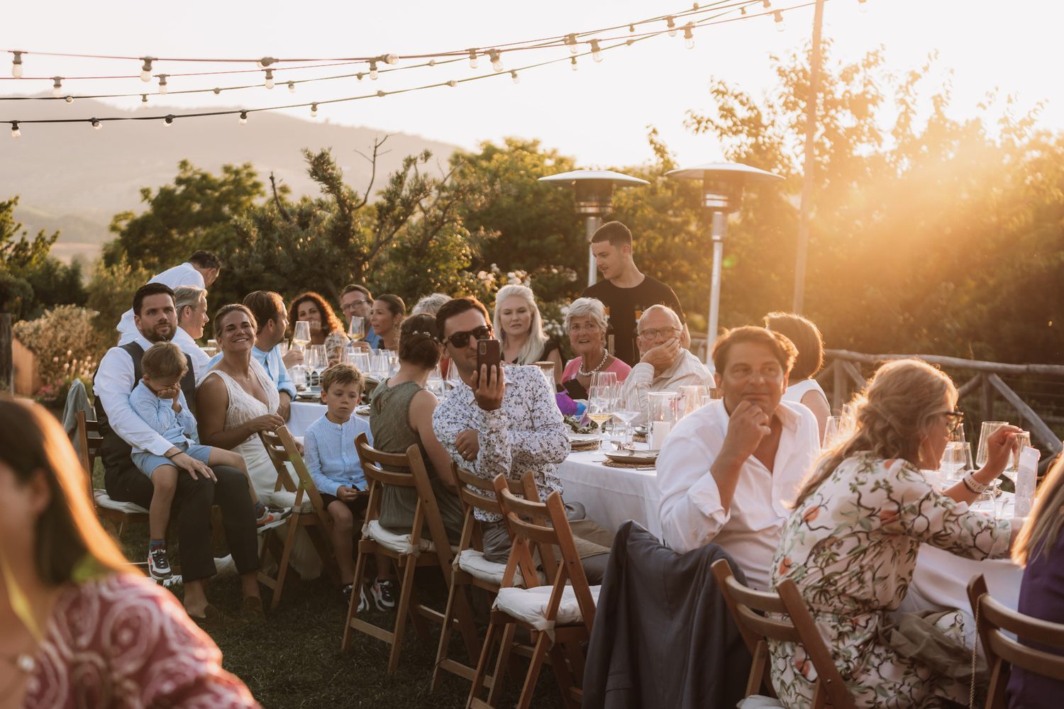 Outdoor dinner party guests seated at long tables during golden hour sunset in a garden setting.