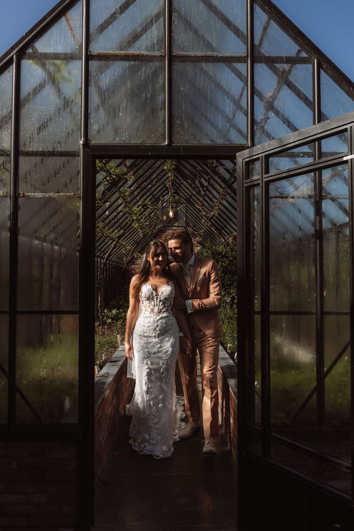 A couple stands together in a moody glass greenhouse wearing formal attire with dramatic lighting.