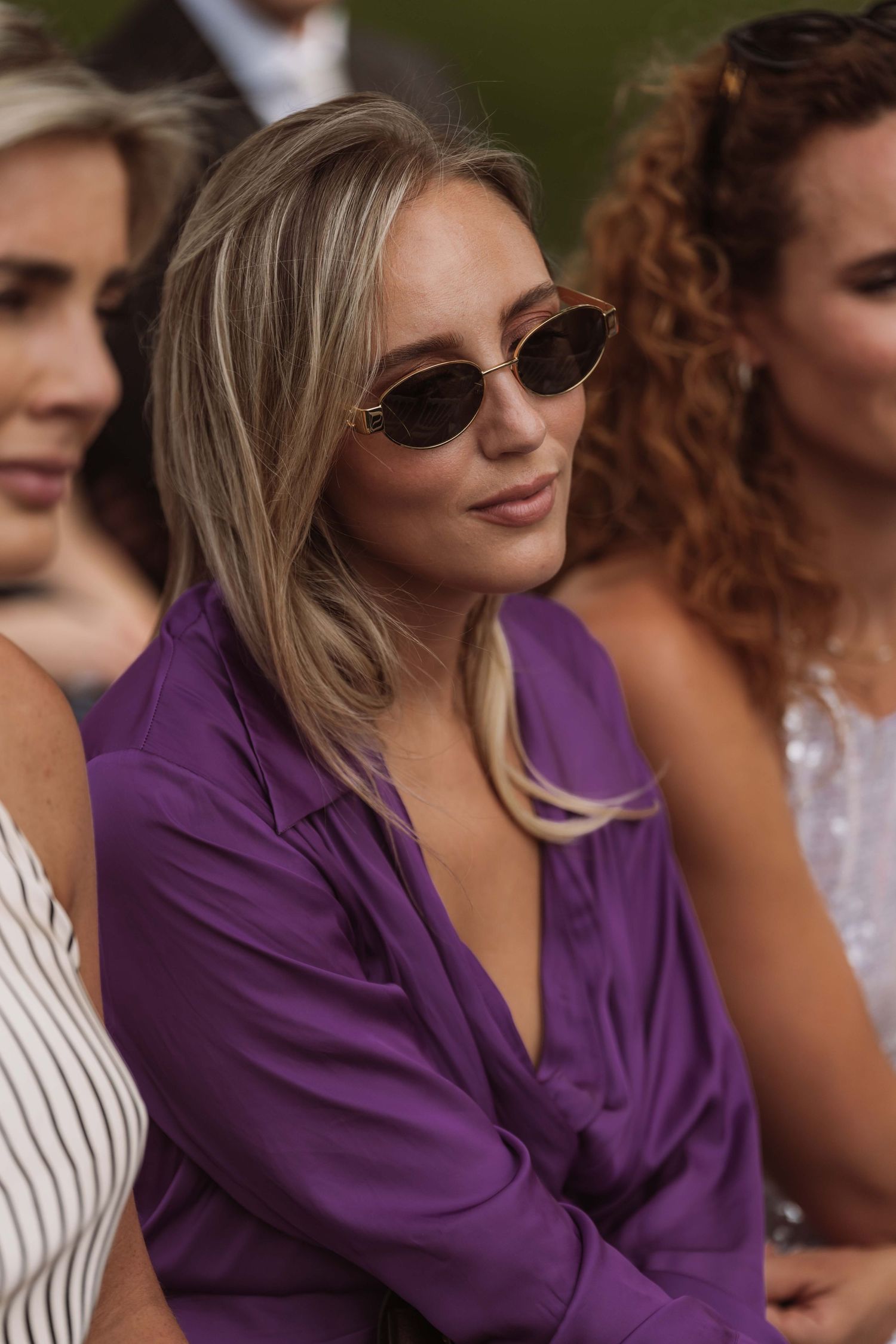 A person in a purple blazer and sunglasses sits outdoors at a social gathering.