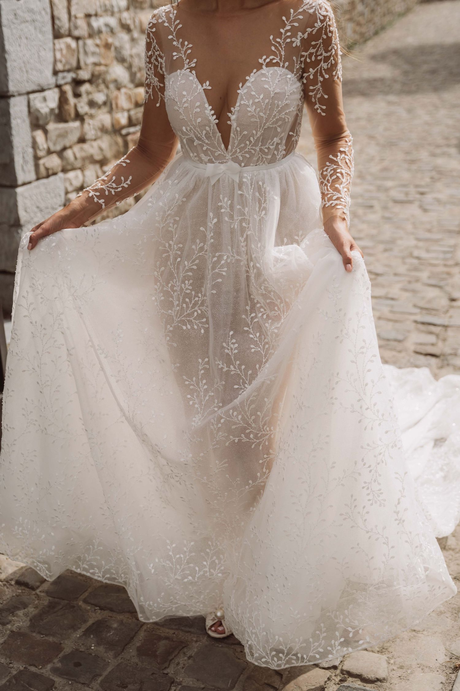 Close-up details of a romantic lace wedding dress with delicate beading and flowing tulle skirt on cobblestone street.