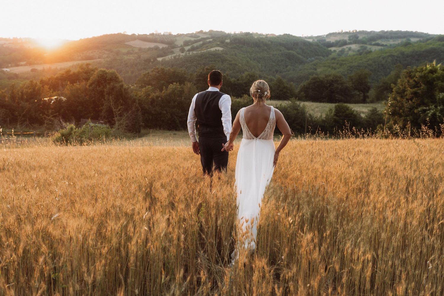 A couple stands in a golden wheat field at sunset with rolling hills and mountains in the background.