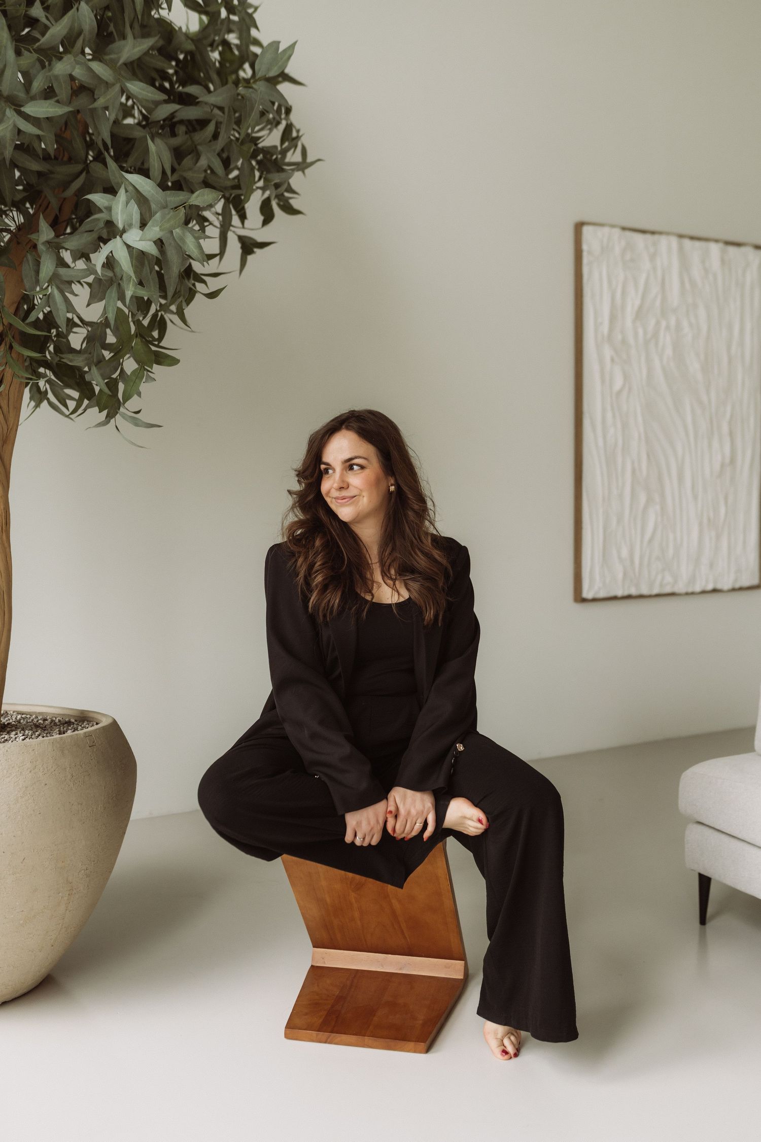 Minimalist interior scene with wooden stool, potted plant, and figure in black outfit sitting against white walls.