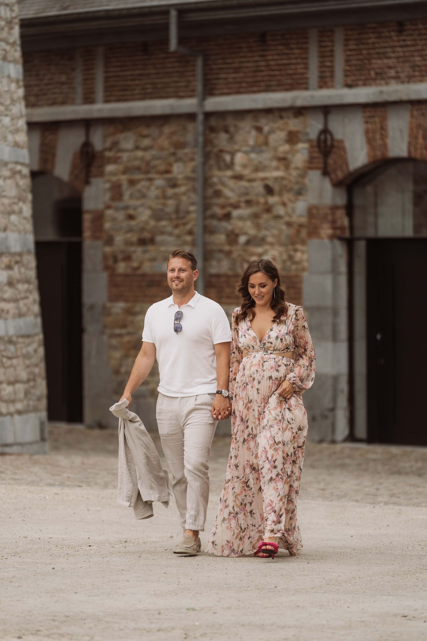 A couple walking together across a rustic stone courtyard, she wearing a floral dress and he in white casual attire.