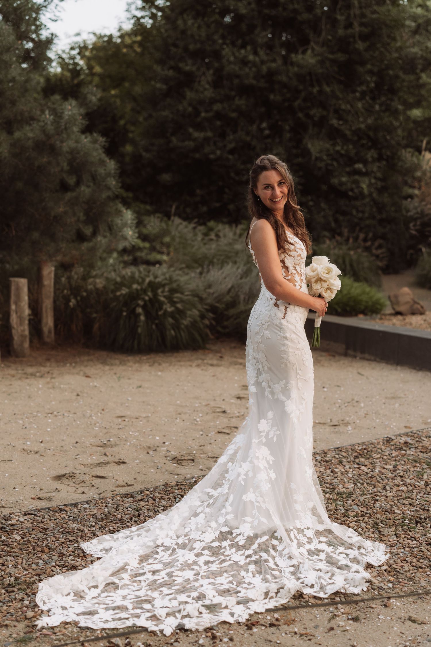 A bride in a fitted lace wedding gown with long train poses outdoors against a natural backdrop holding a white bouquet.