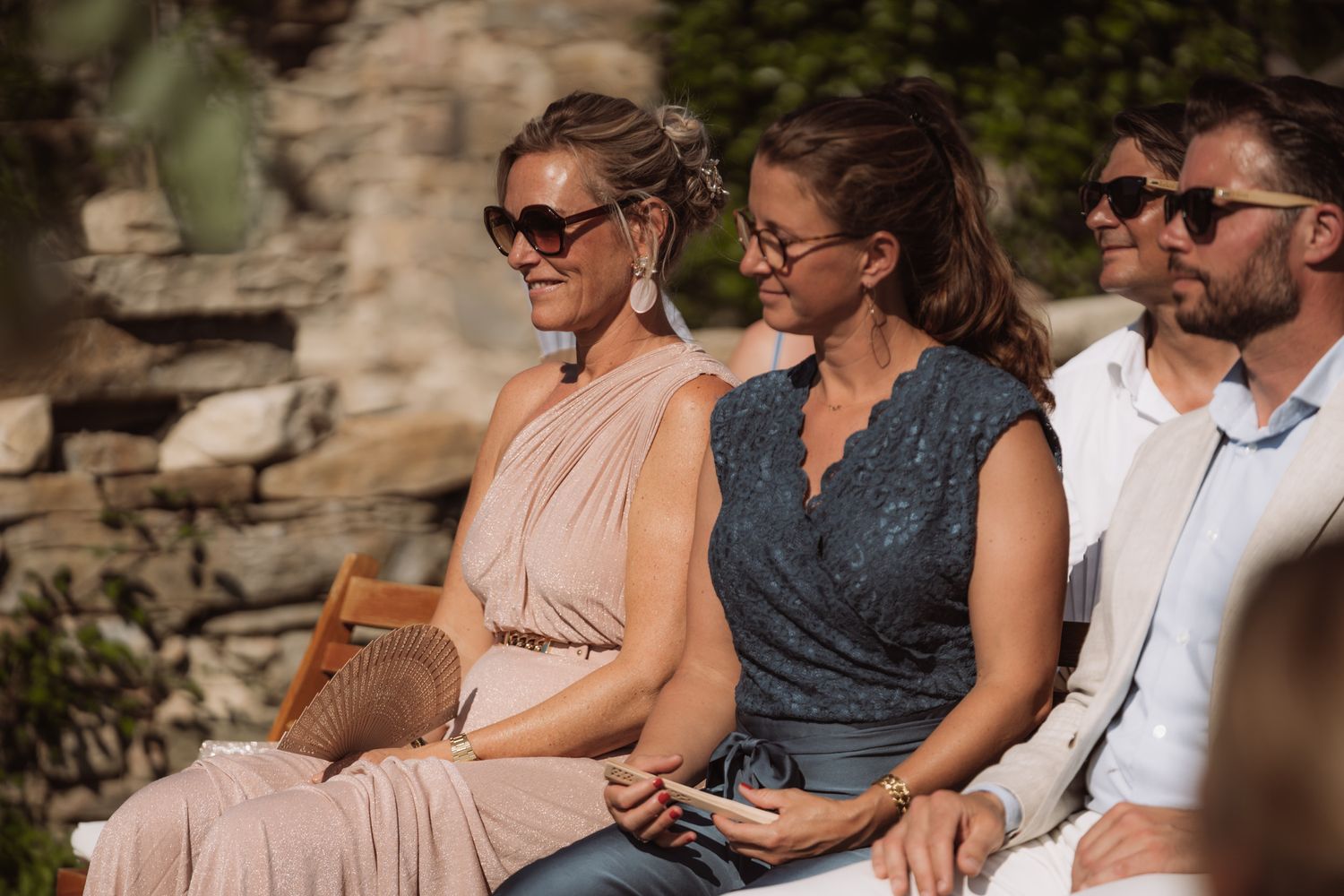 A group of people sit together outdoors laughing and smiling while enjoying a sunny day against a stone wall backdrop.