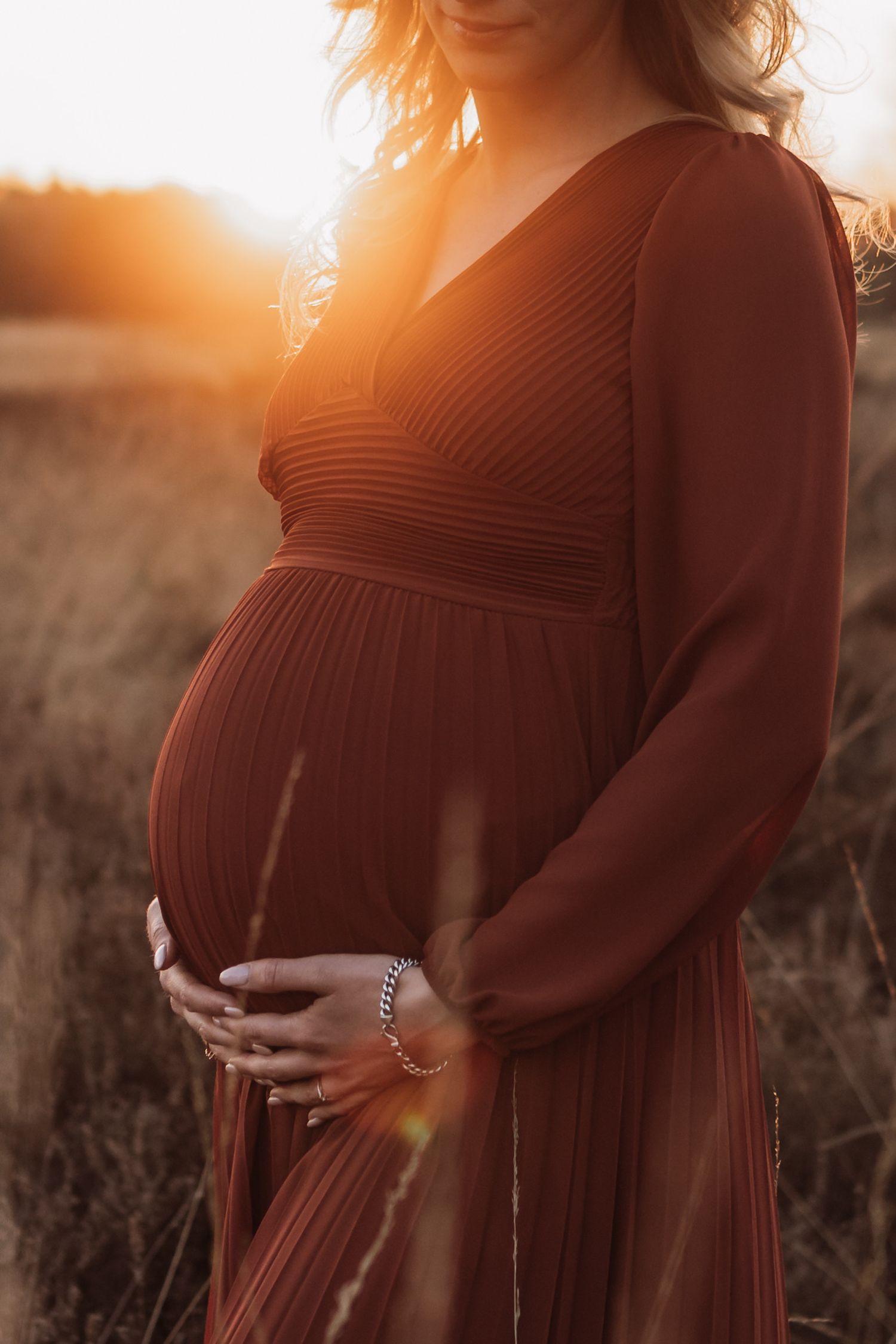 Sunset maternity photos showing a pregnant woman in a rust-colored dress in a field.