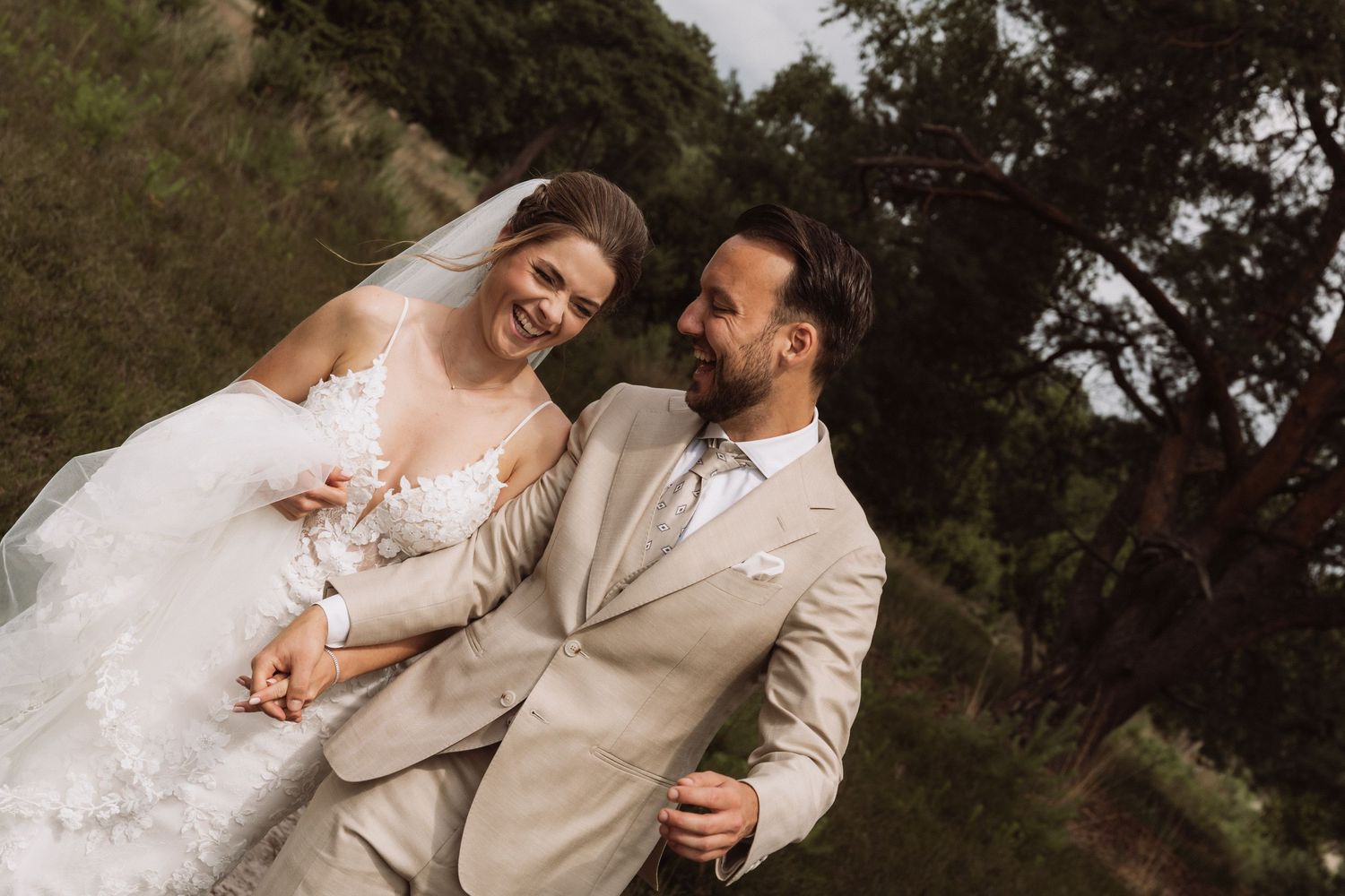 A joyful wedding couple laughs together in a natural outdoor setting.