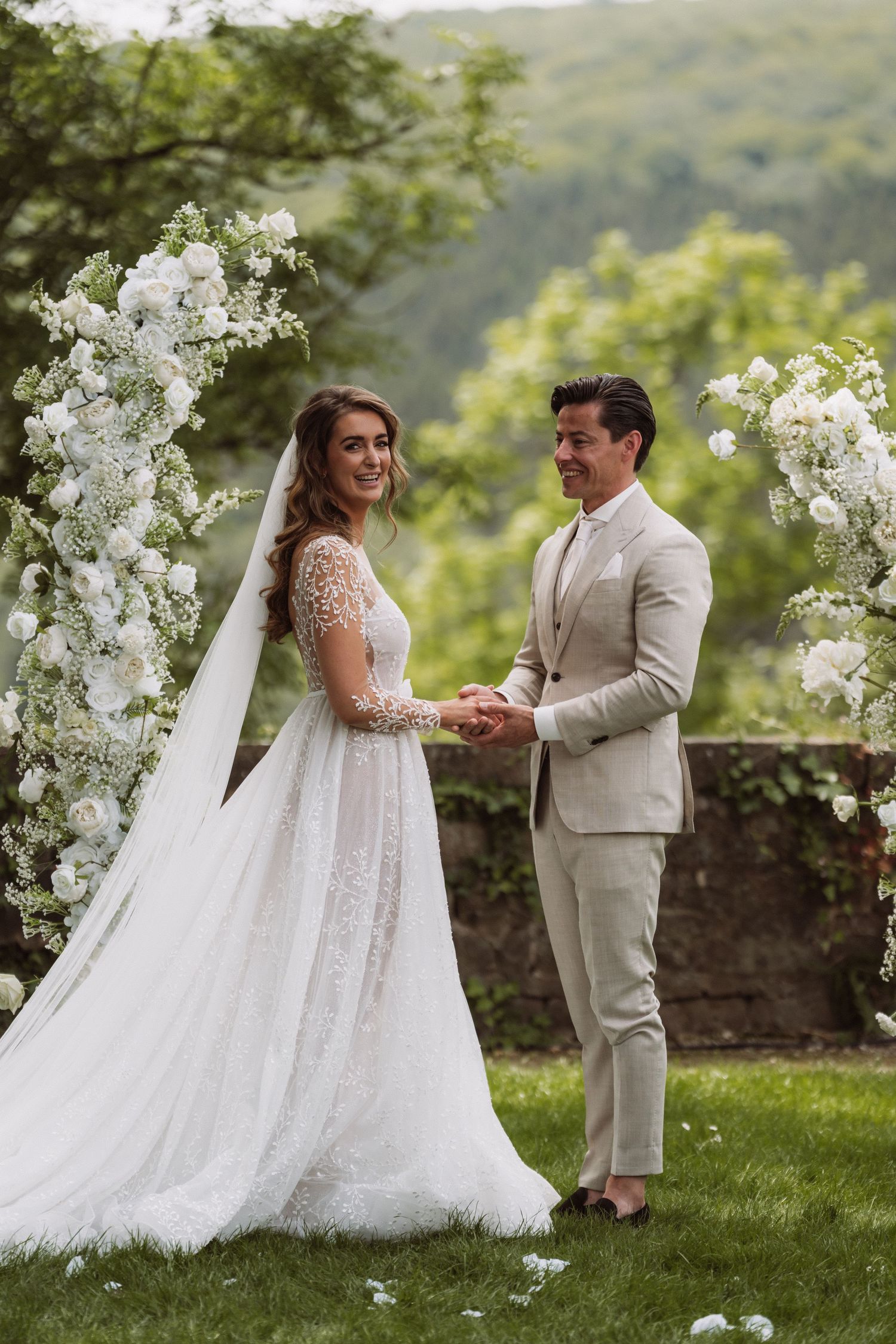 A wedding couple stands together under white floral arches in a scenic mountain setting.