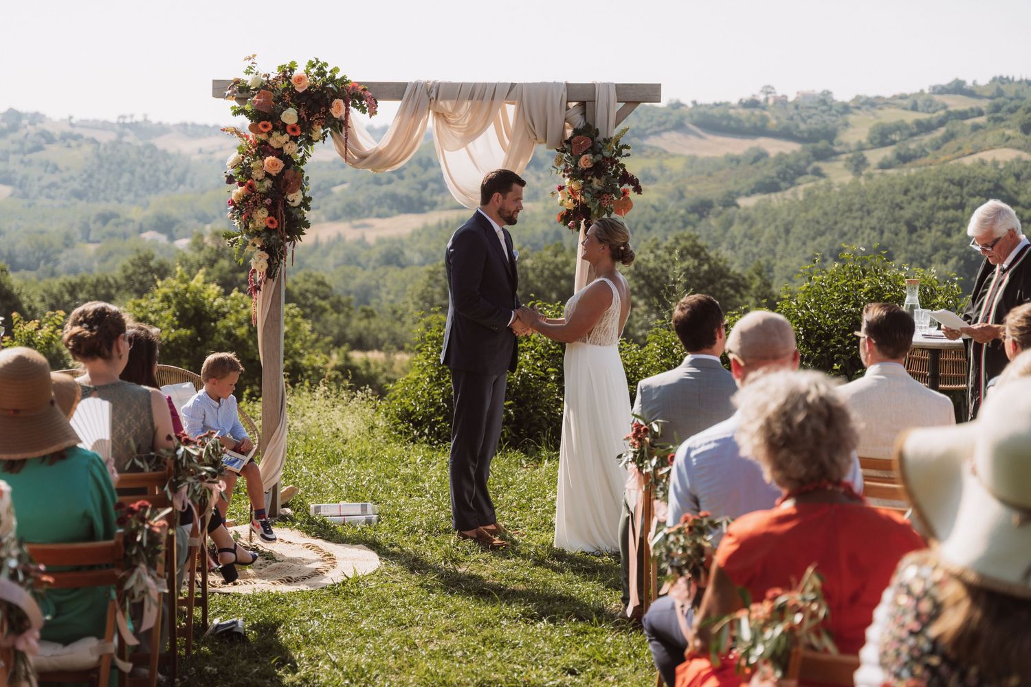A romantic outdoor wedding ceremony overlooking rolling hills with autumn floral decorations and guests seated on chairs.
