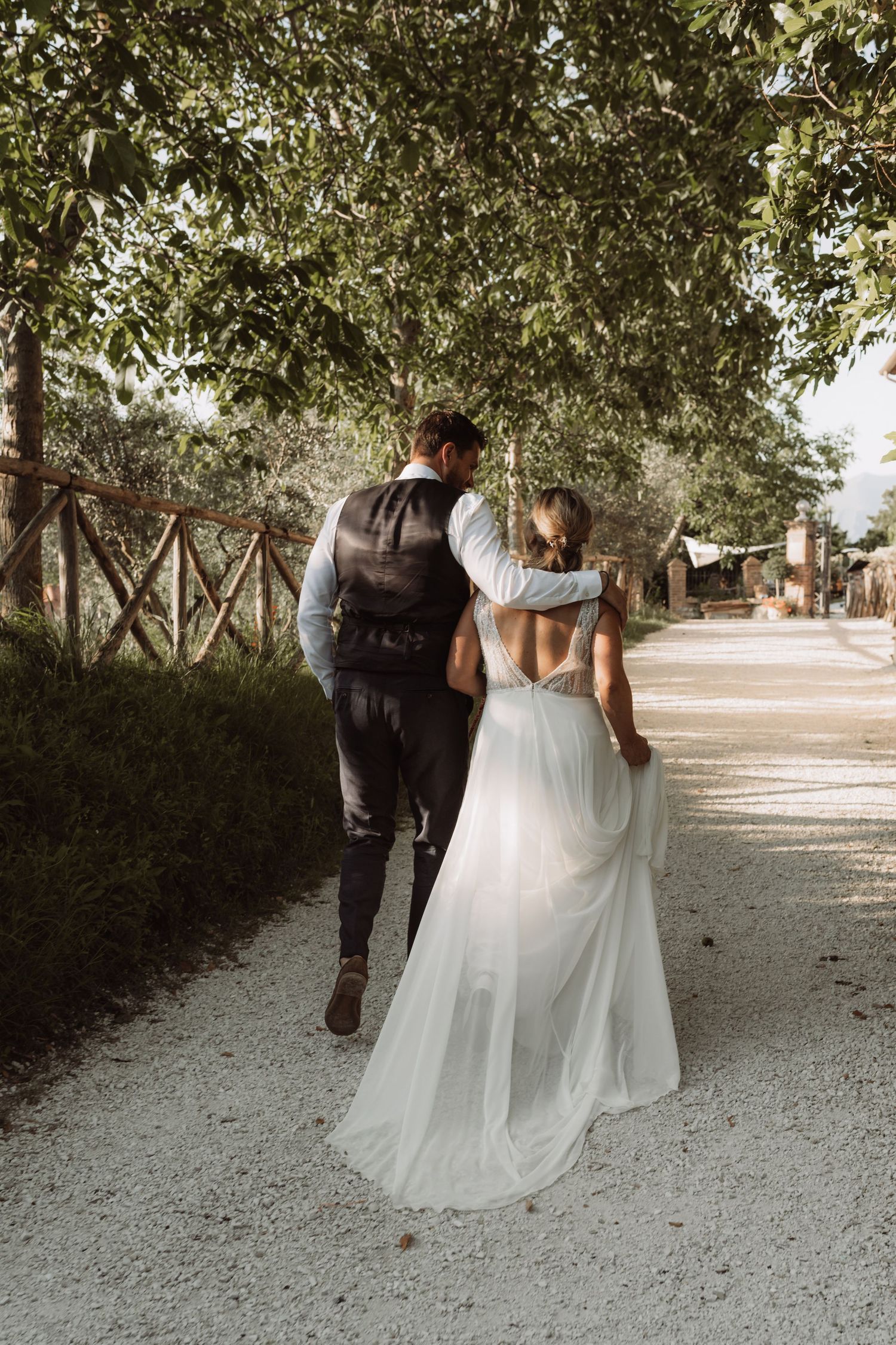 A romantic wedding couple walking down a tree-lined path, with the bride wearing a flowing white dress.