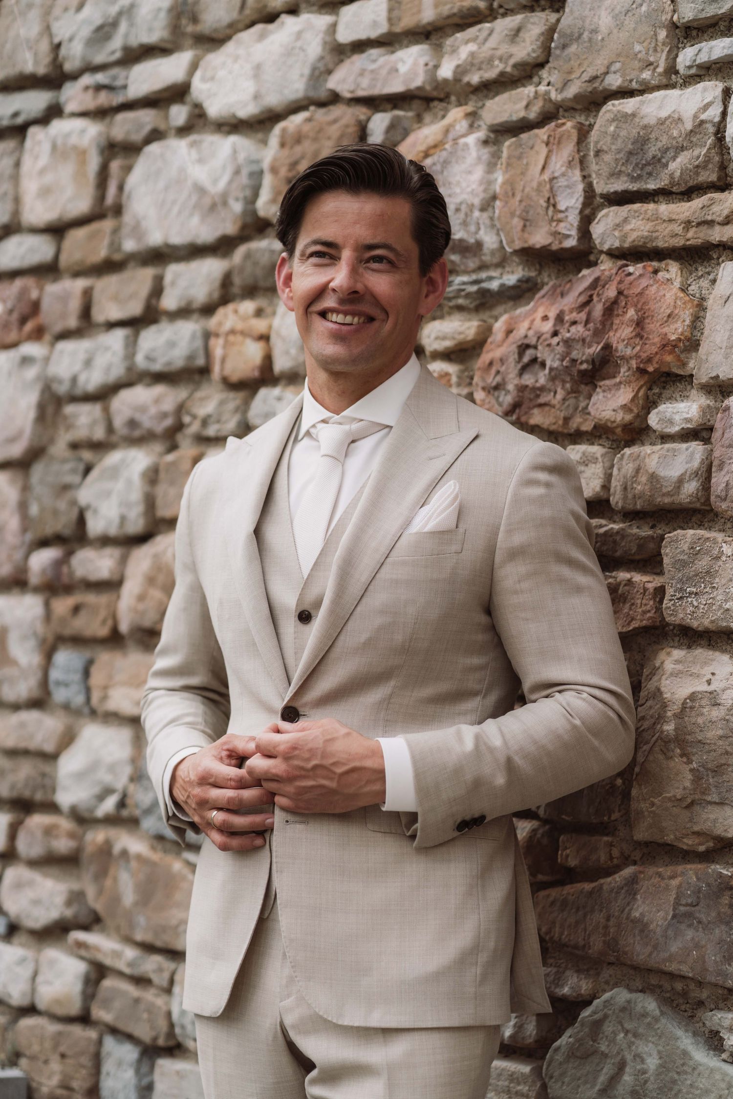 A groom in a light beige suit stands against a rustic stone wall backdrop during a wedding photoshoot.