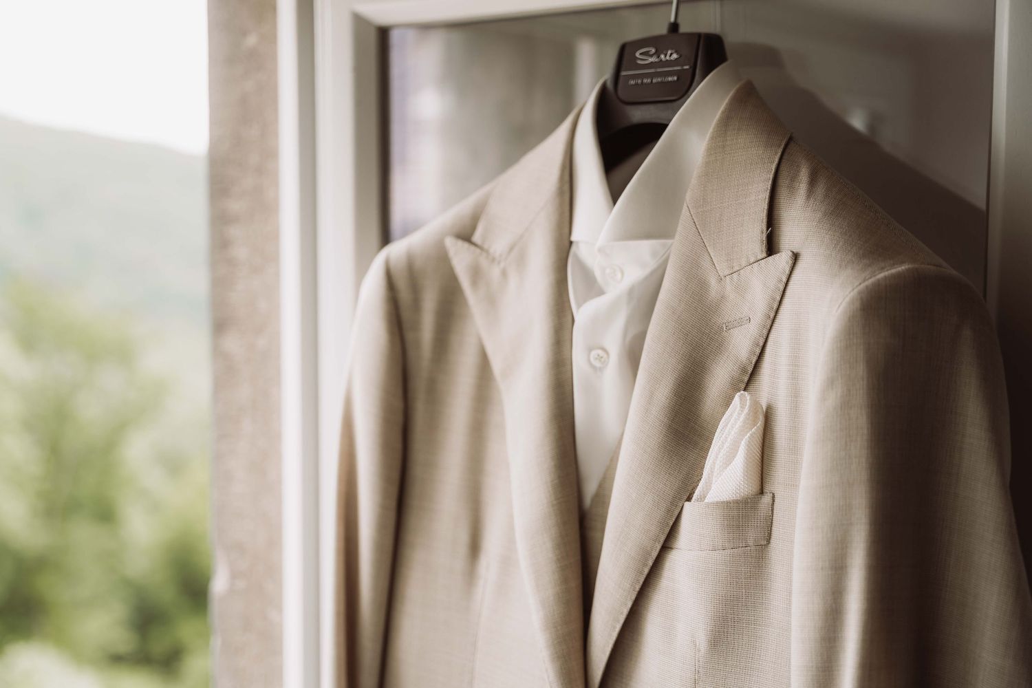 A light beige wedding suit jacket hangs on a wooden hanger by a window with natural lighting.
