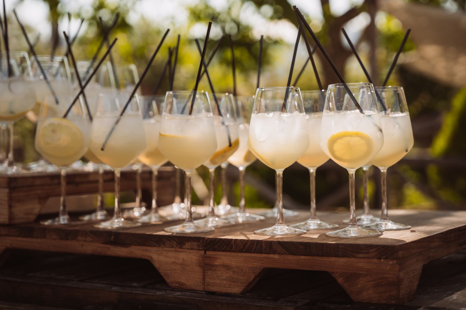 A rustic wooden tray holds elegant wine glasses on an outdoor table setting with natural lighting and blurred background.