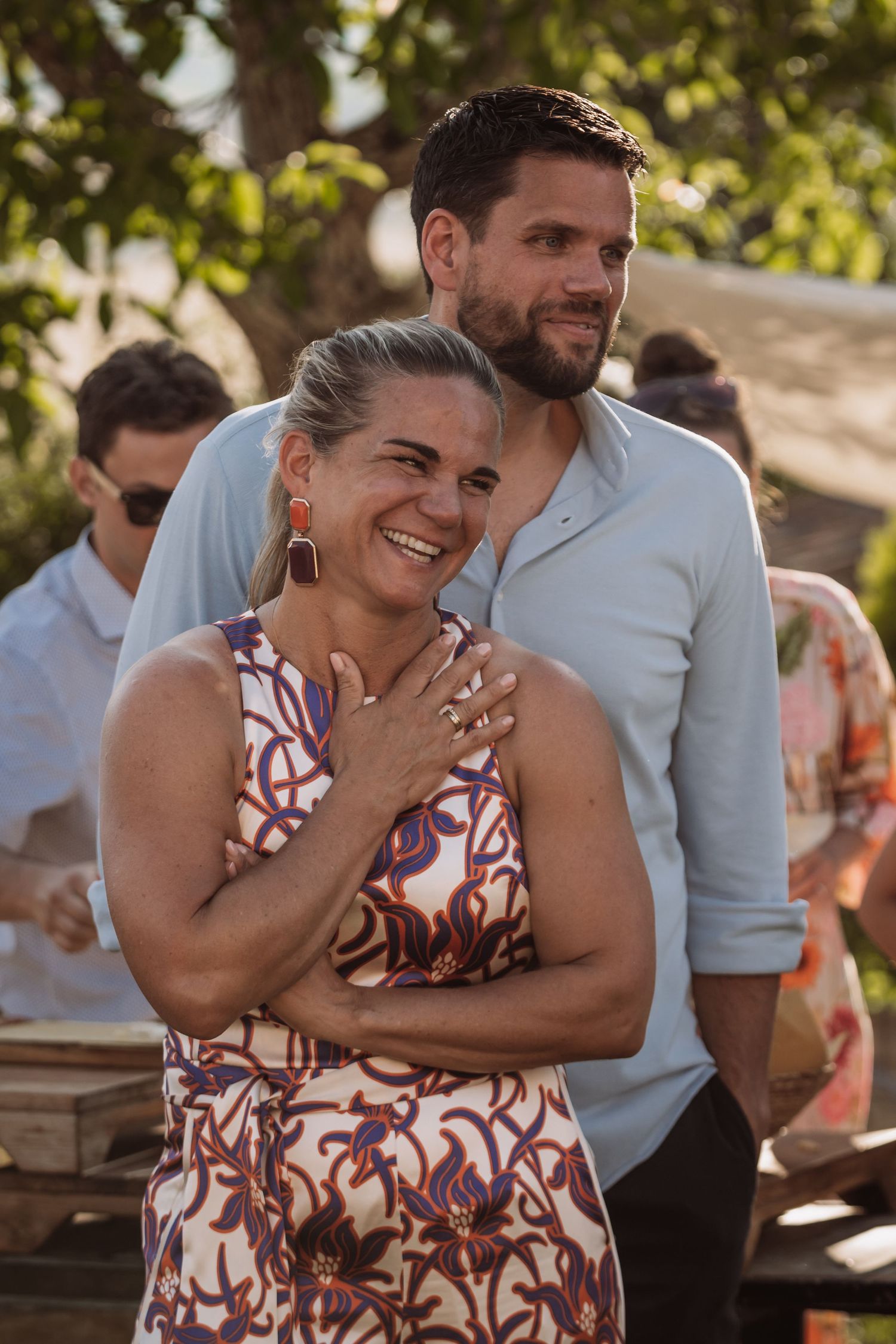 A couple shares a joyful moment together at an outdoor gathering, laughing and embracing in natural sunlight.