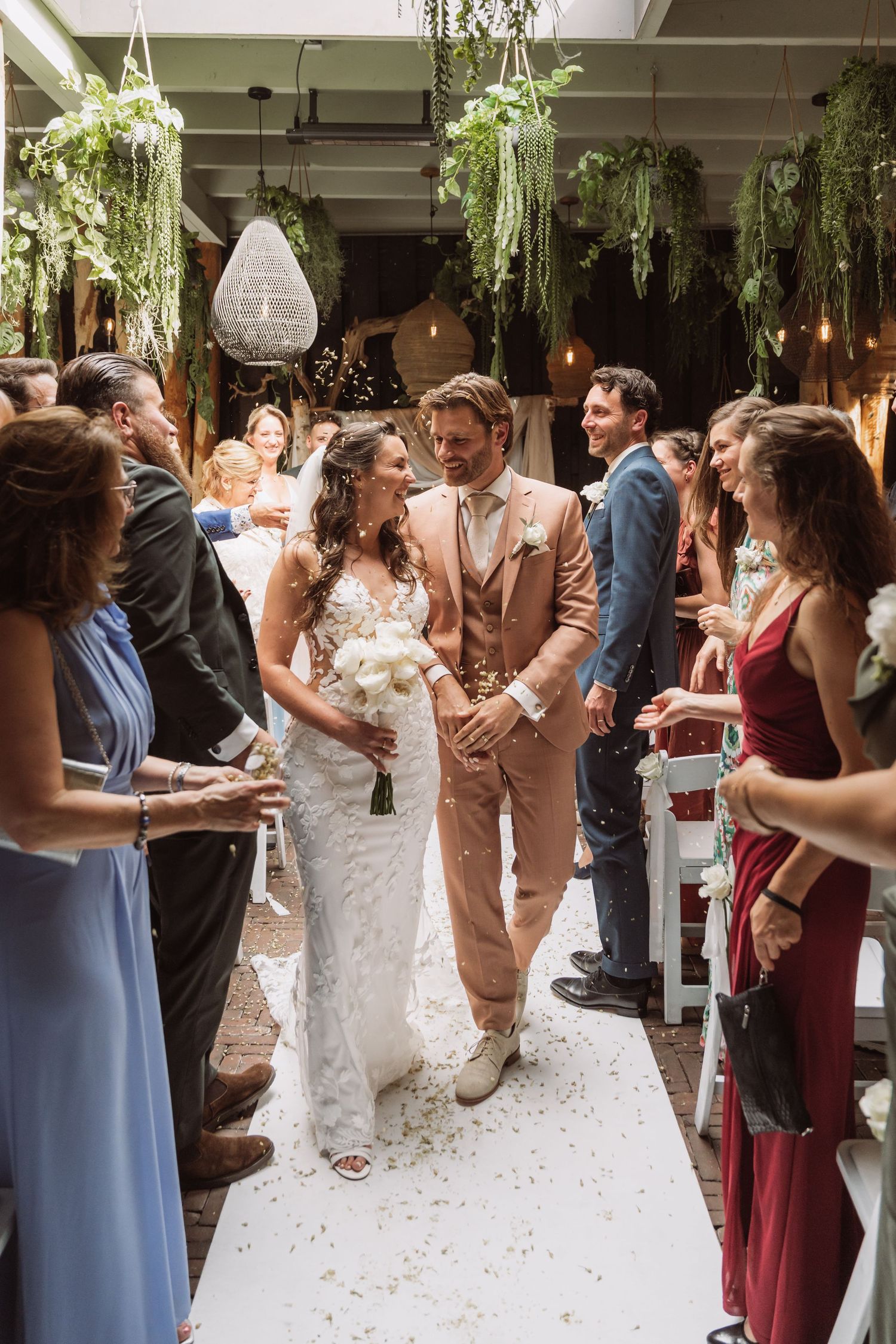 A wedding ceremony in a venue decorated with hanging plants as guests line the aisle while flower petals scatter the floor.