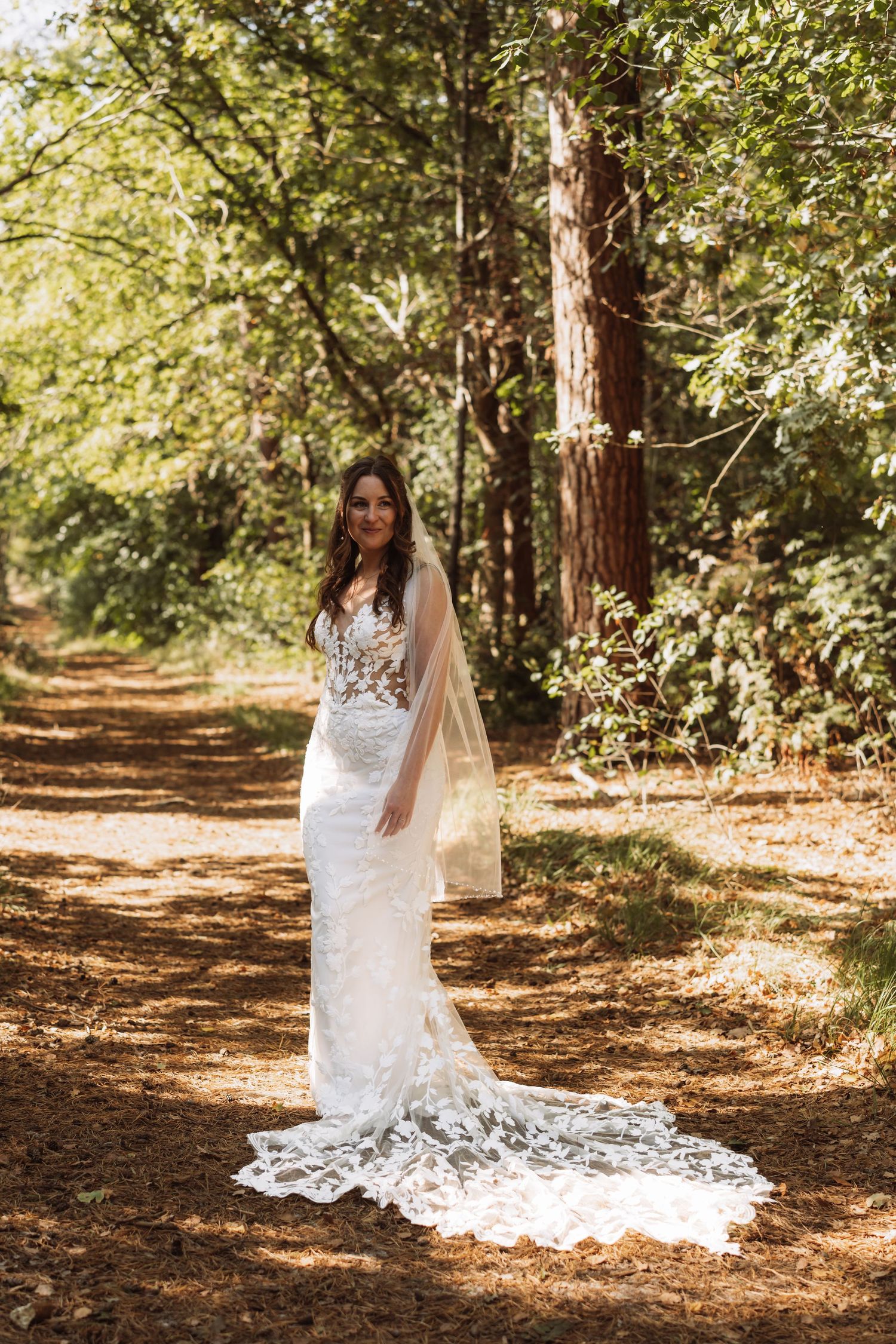 A bride in a white lace wedding dress with long train poses on a sunlit forest path surrounded by tall trees.