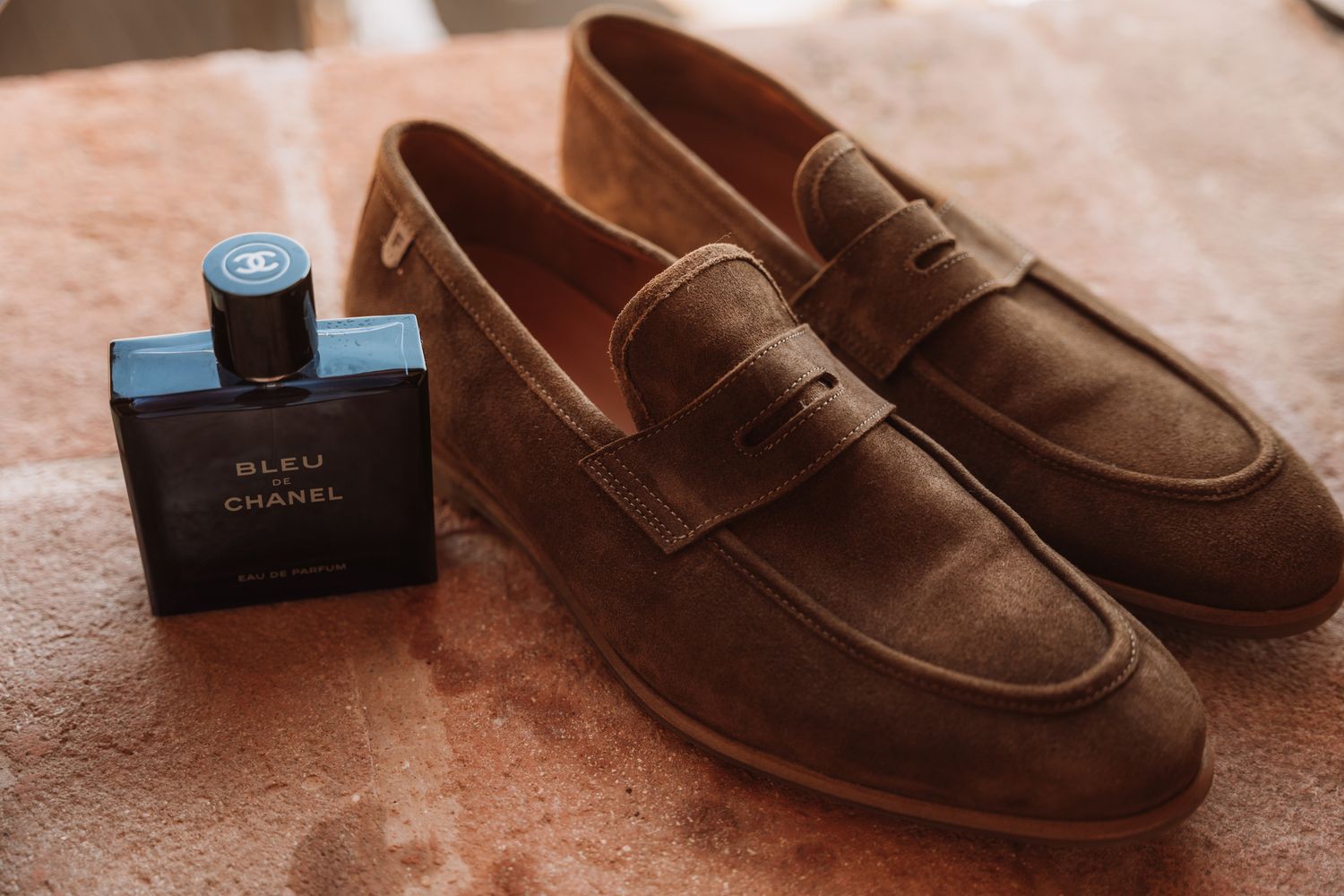 Close-up view of brown suede penny loafers with a blue perfume bottle displayed on a textured surface.